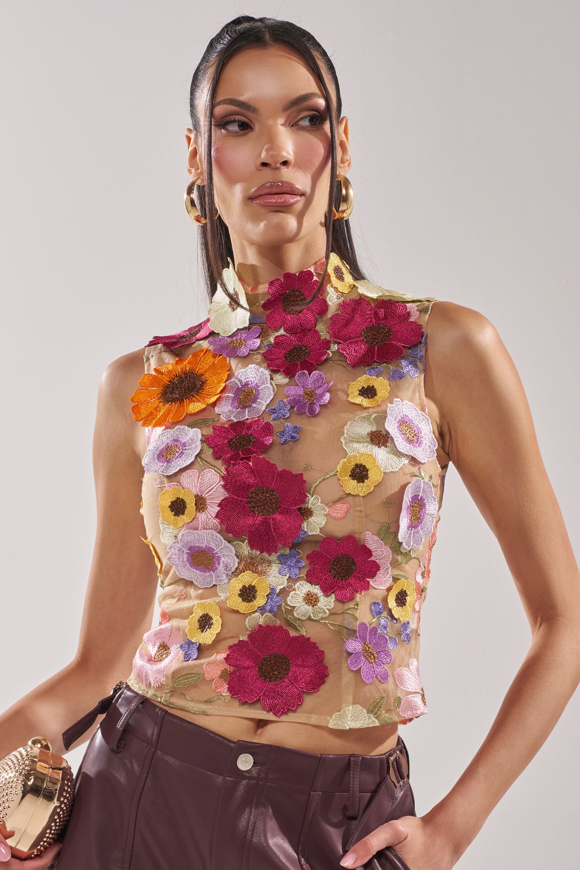 A woman with long dark hair wears the WHERE THIS FLOWER BLOOMS MESH TOP, a sleeveless, fitted sheer mesh piece with colorful floral embroidery. She pairs it with dark high-waisted pants and gold hoop earrings. 1