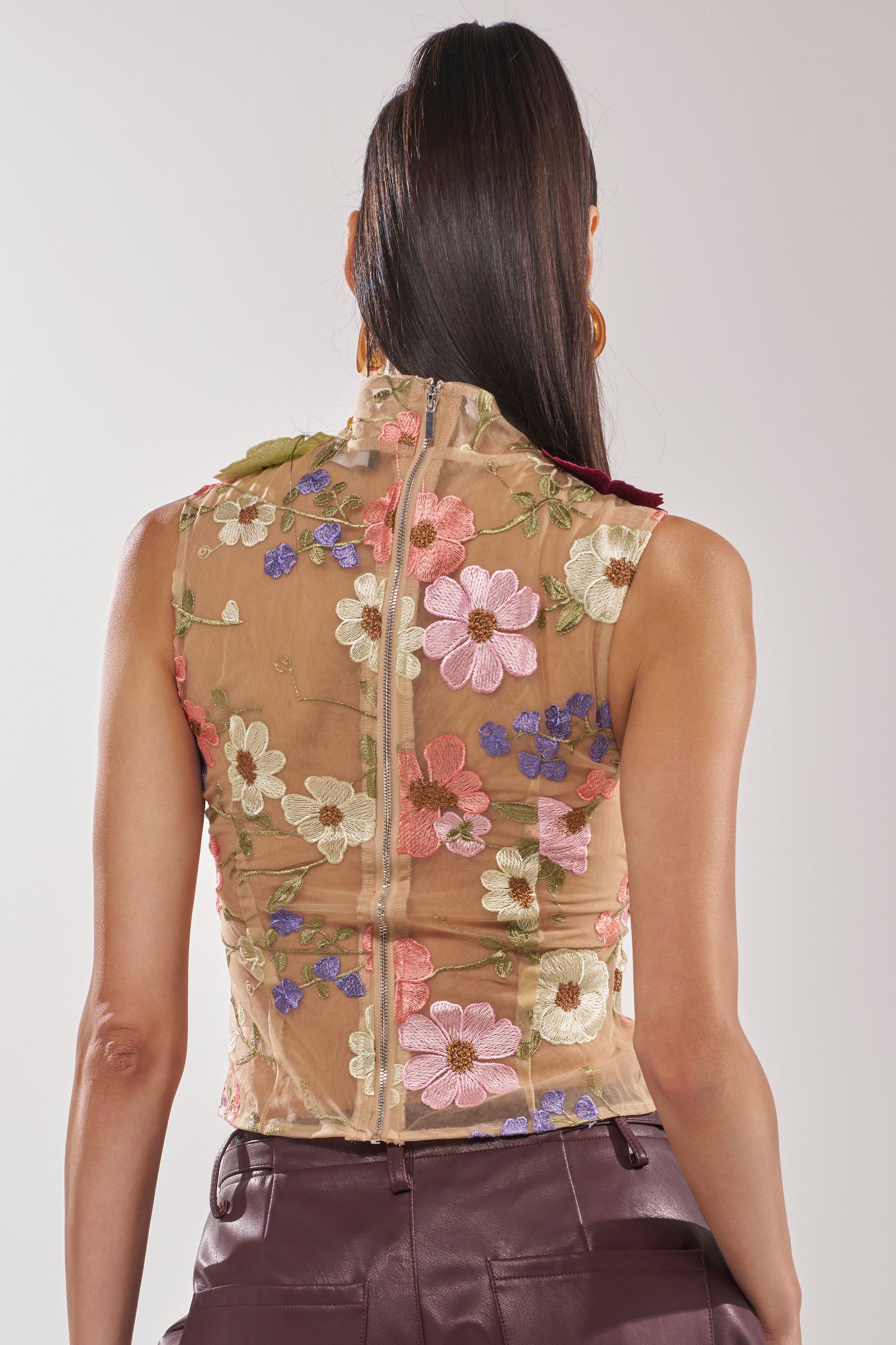 A woman with long brown hair is seen from the back against a plain background, wearing the WHERE THIS FLOWER BLOOMS MESH TOP featuring sheer mesh with 3D embroidered floral appliqués and paired with dark high-waisted pants. 3