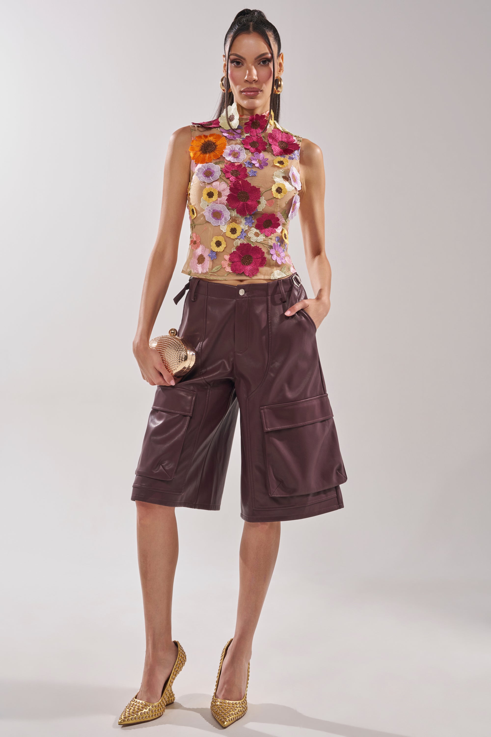 A woman models the WHERE THIS FLOWER BLOOMS MESH TOP, featuring a sheer mesh design with colorful 3D floral appliqués, styled with burgundy leather knee-length shorts, gold studded heels, and gold accessories against a plain background. 4
