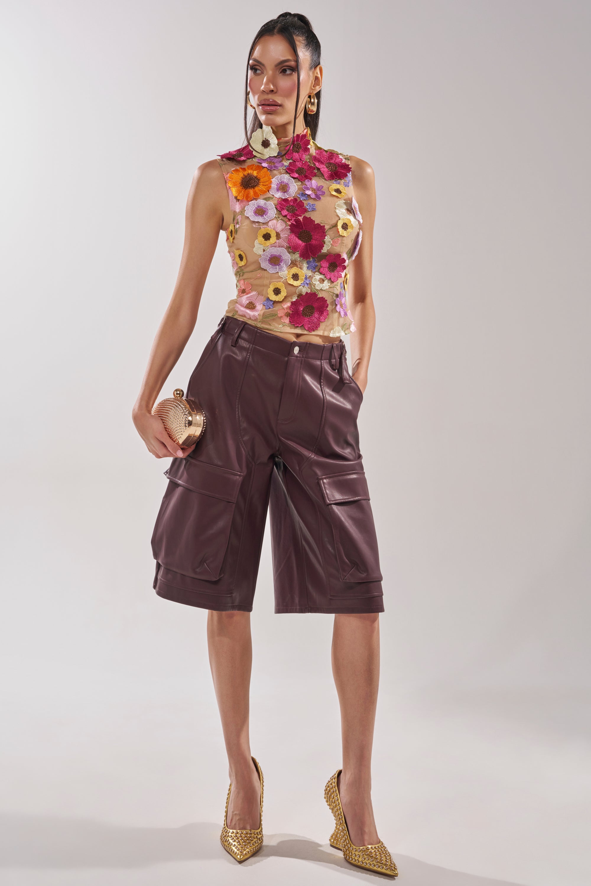 A woman poses in the WHERE THIS FLOWER BLOOMS MESH TOP paired with maroon leather shorts, gold high heels, a glittery clutch, and bold earrings. Her hair is styled half up, half down. 5