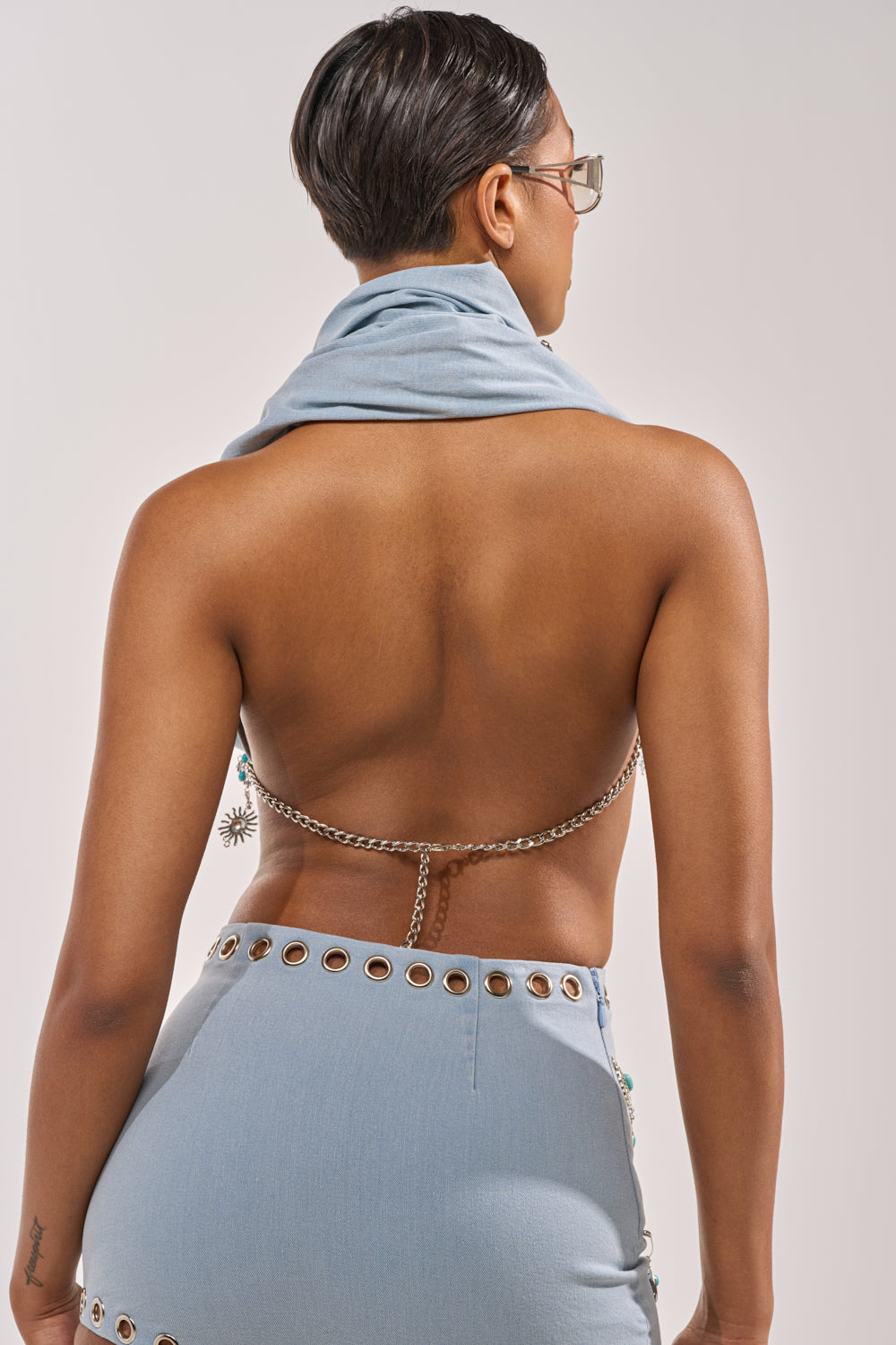 A person with short hair and glasses is seen from behind wearing the FESTIVAL GIRLY COWL NECK BLOUSE, a backless light blue top with chain straps, paired with a matching skirt and draped scarf. 3