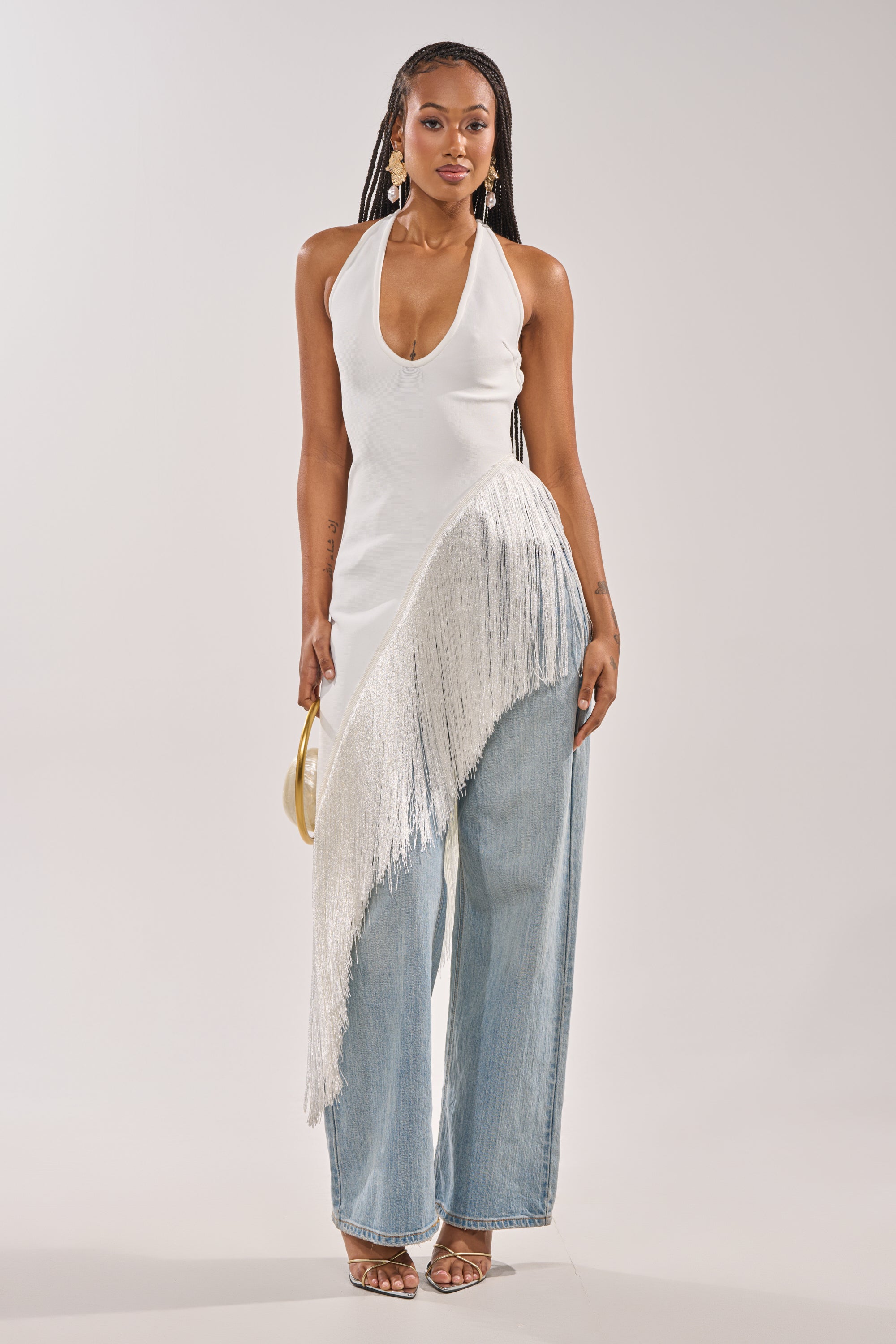 A woman wears the TAKE ME OUT FRINGE TOP—white with silver diagonal fringe—paired with wide-leg light blue jeans, gold sandals, and a small gold handbag for a chic evening look. 2