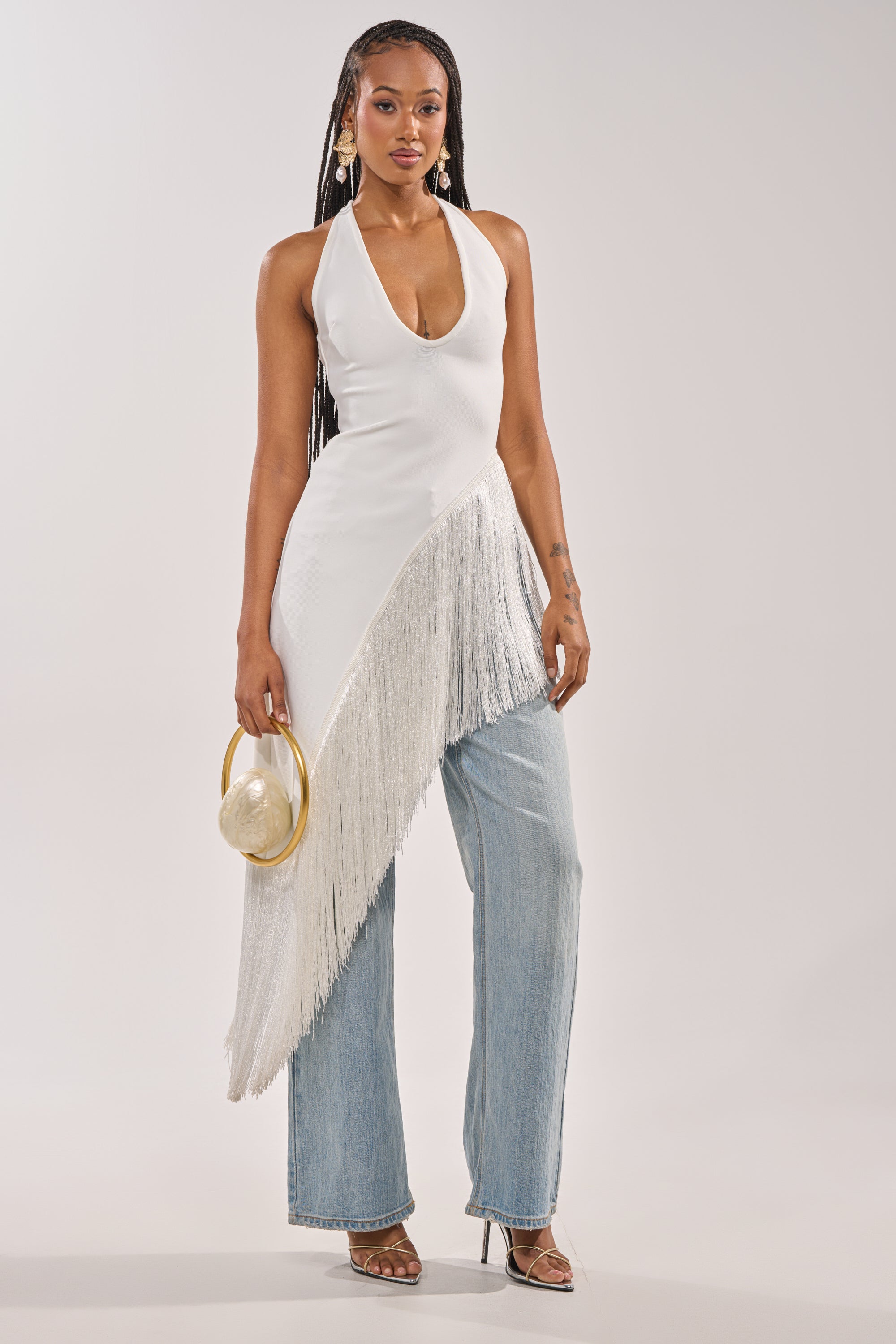 A woman models the TAKE ME OUT FRINGE TOP—a white halter paired with light blue jeans, transparent heels, and a round handbag. With long braids and statement earrings, she stands against a plain background, ideal for an evening occasion. 3