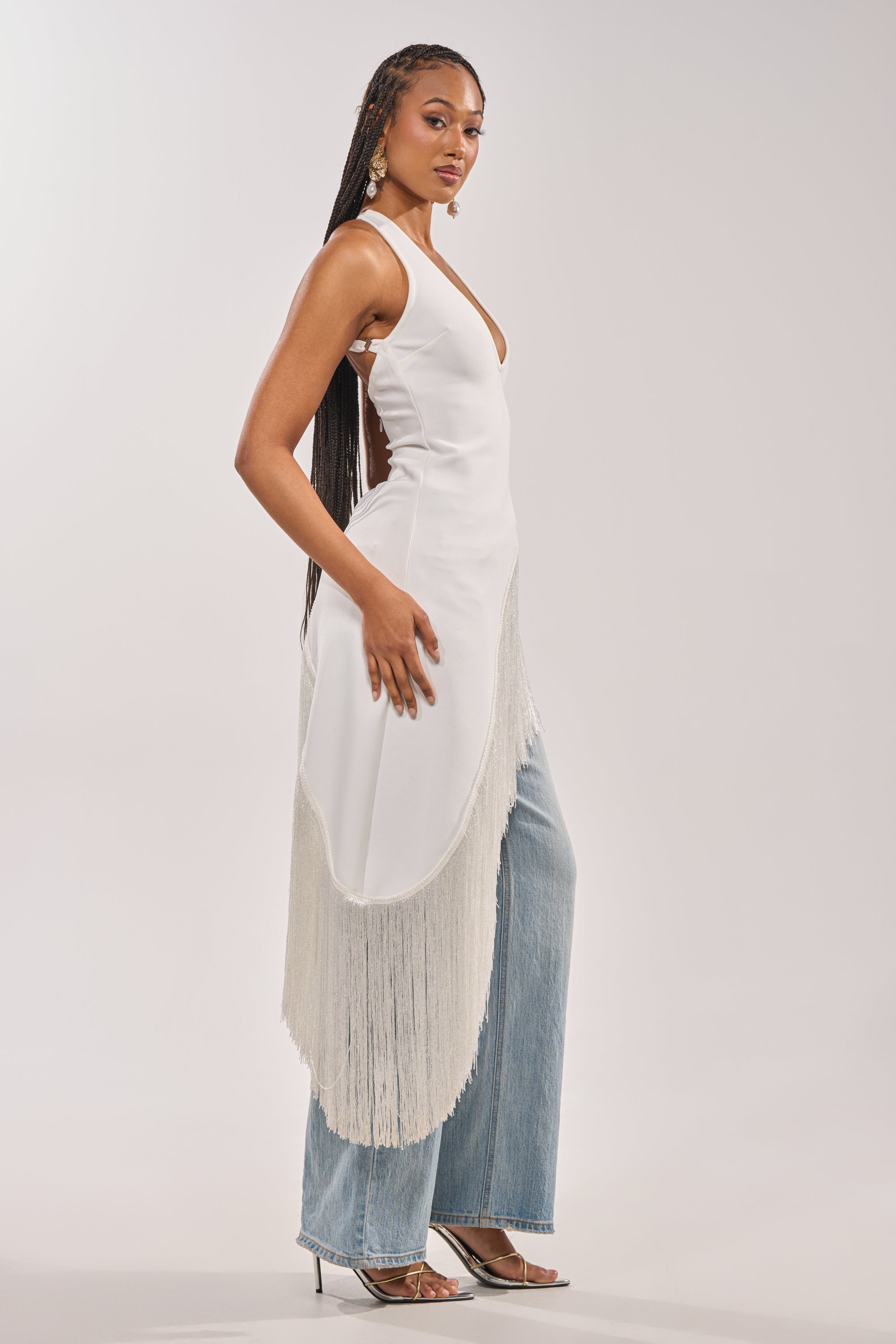 A woman with long braids wears the TAKE ME OUT FRINGE TOP—a white halter style with an asymmetrical hem—over light blue wide-leg jeans and silver heels, posing sideways against a light neutral background for an effortlessly chic look. 4