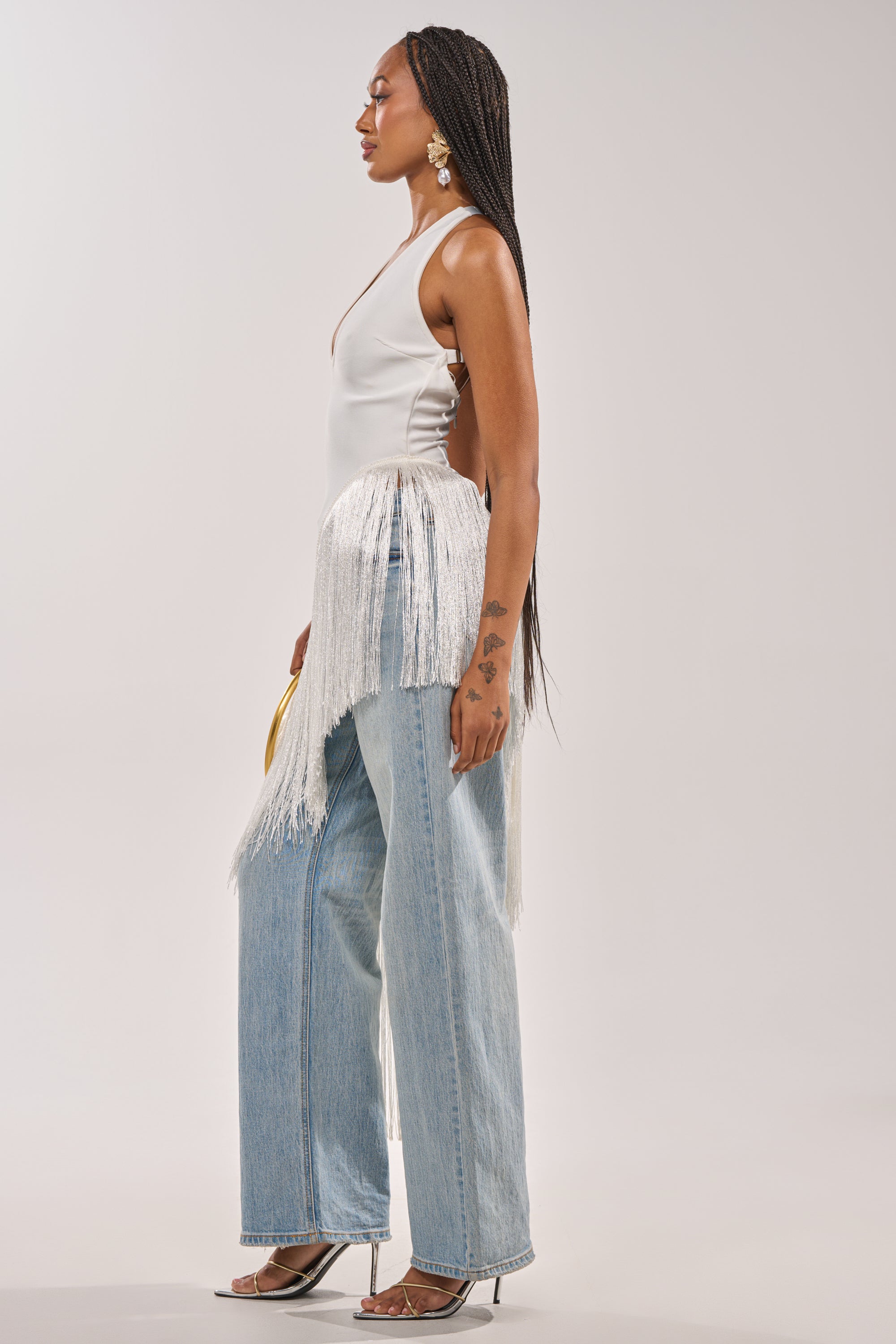 A woman stands in profile wearing the TAKE ME OUT FRINGE TOP, light blue wide-leg jeans, clear heels, and large earrings. With long braided hair and visible arm tattoos, she creates an effortless evening statement look. 5