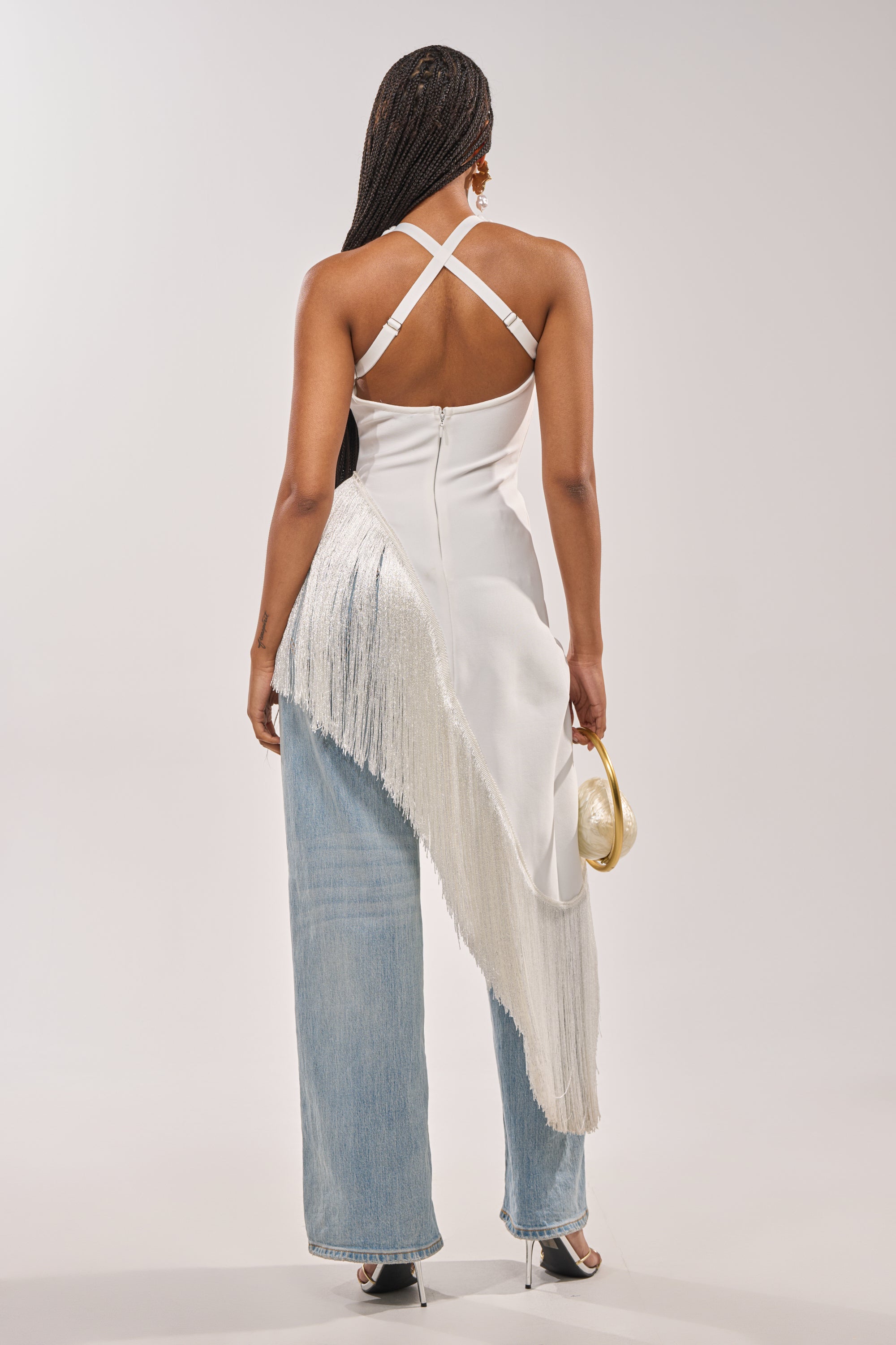 A woman with long braids stands with her back to the camera in the TAKE ME OUT FRINGE TOP, paired with light blue jeans, black heels, and a small gold clutch—a chic evening statement look. 6