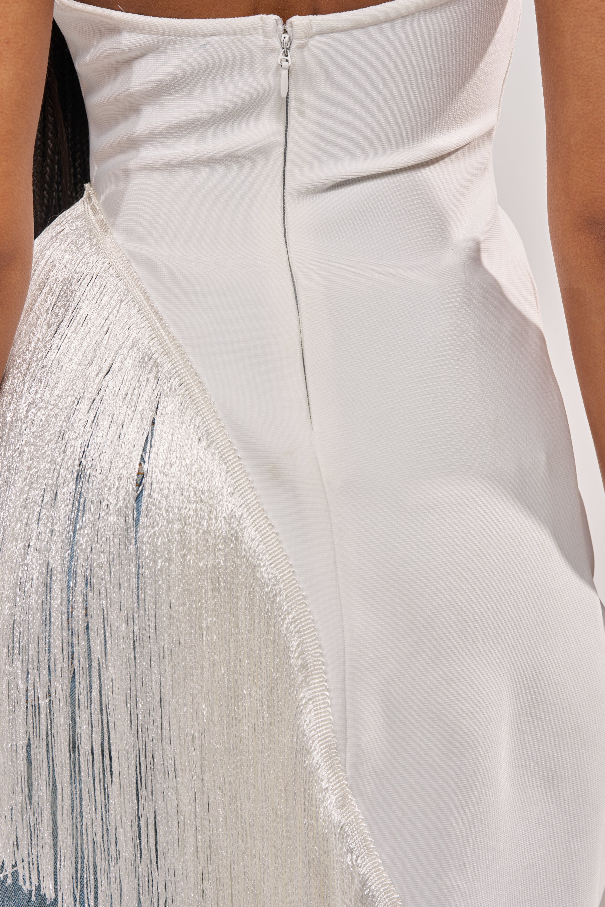Close-up of the TAKE ME OUT FRINGE TOP: a white fitted piece featuring a back zipper and diagonal row of long, shimmering fringe. Its smooth fabric and bold fringe create a sophisticated, modern statement look. 7