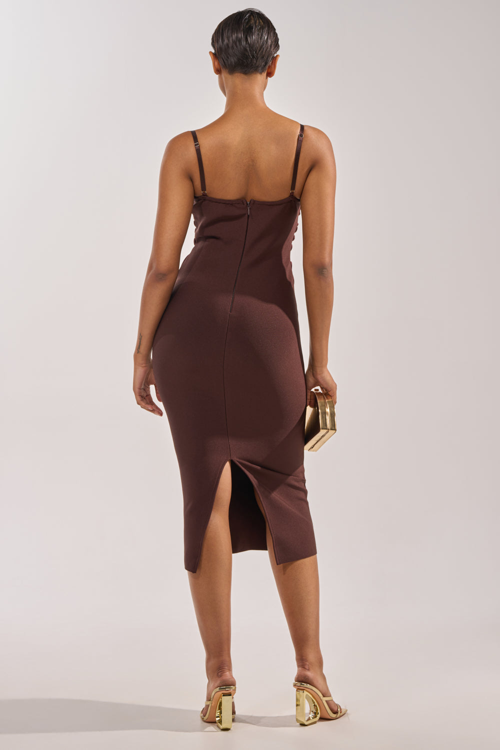 A person with short hair stands with their back to the camera, wearing the ANYTHING BUT BASIC MAXI DRESS in brown, featuring thin straps, a sheer mesh bodice, and back slit, paired with a gold clutch and gold heeled sandals. 3