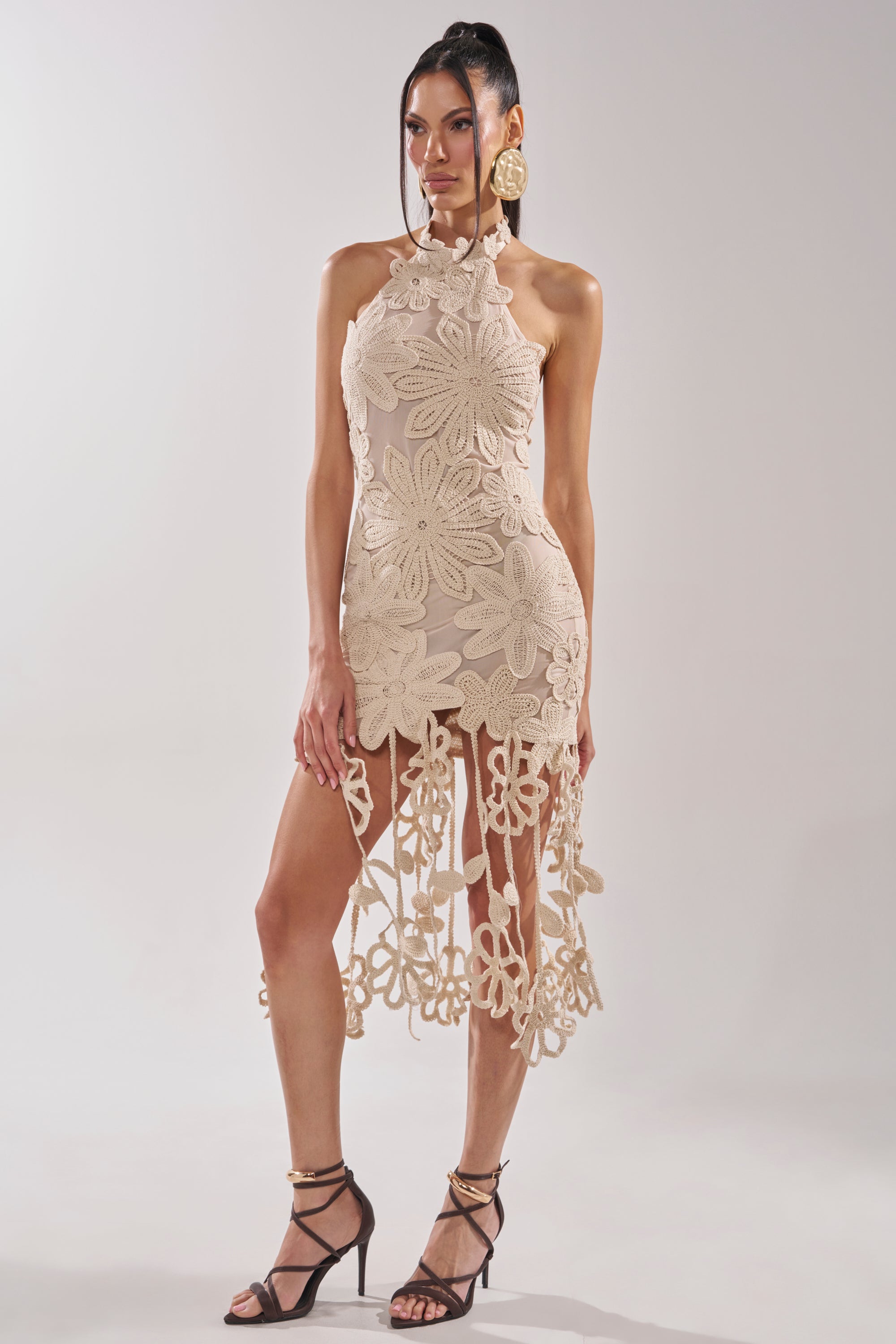 A woman poses in the CELINE FLORAL MIDI DRESS, an ivory crochet lace midi with large floral patterns and fringe details, styled with black strappy heels and circular earrings against a plain light background. 0