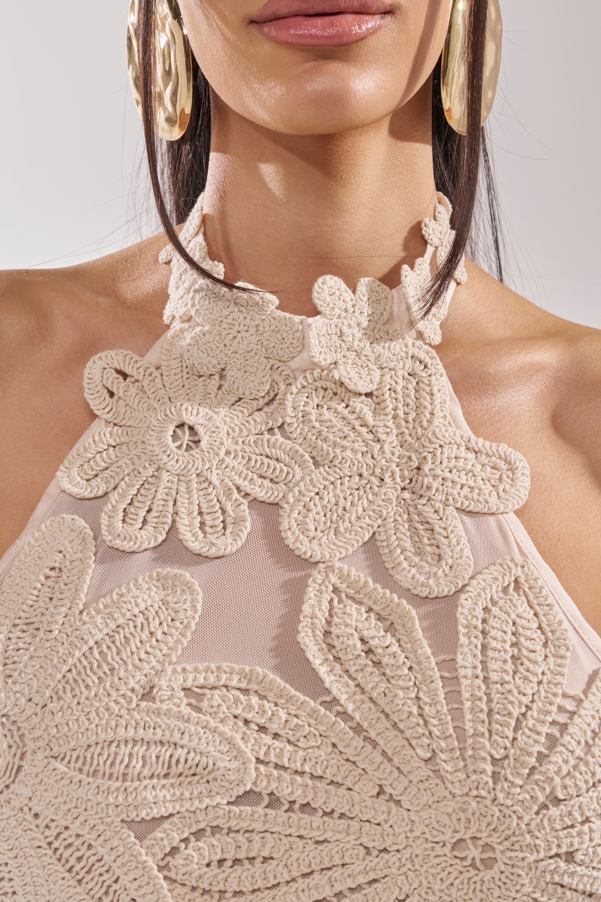 A woman wears large gold earrings and a sheer, nude halter top with cream crochet flowers, echoing the look of the CELINE FLORAL MIDI DRESS. Only her lower face, neck, and shoulders are visible. 9