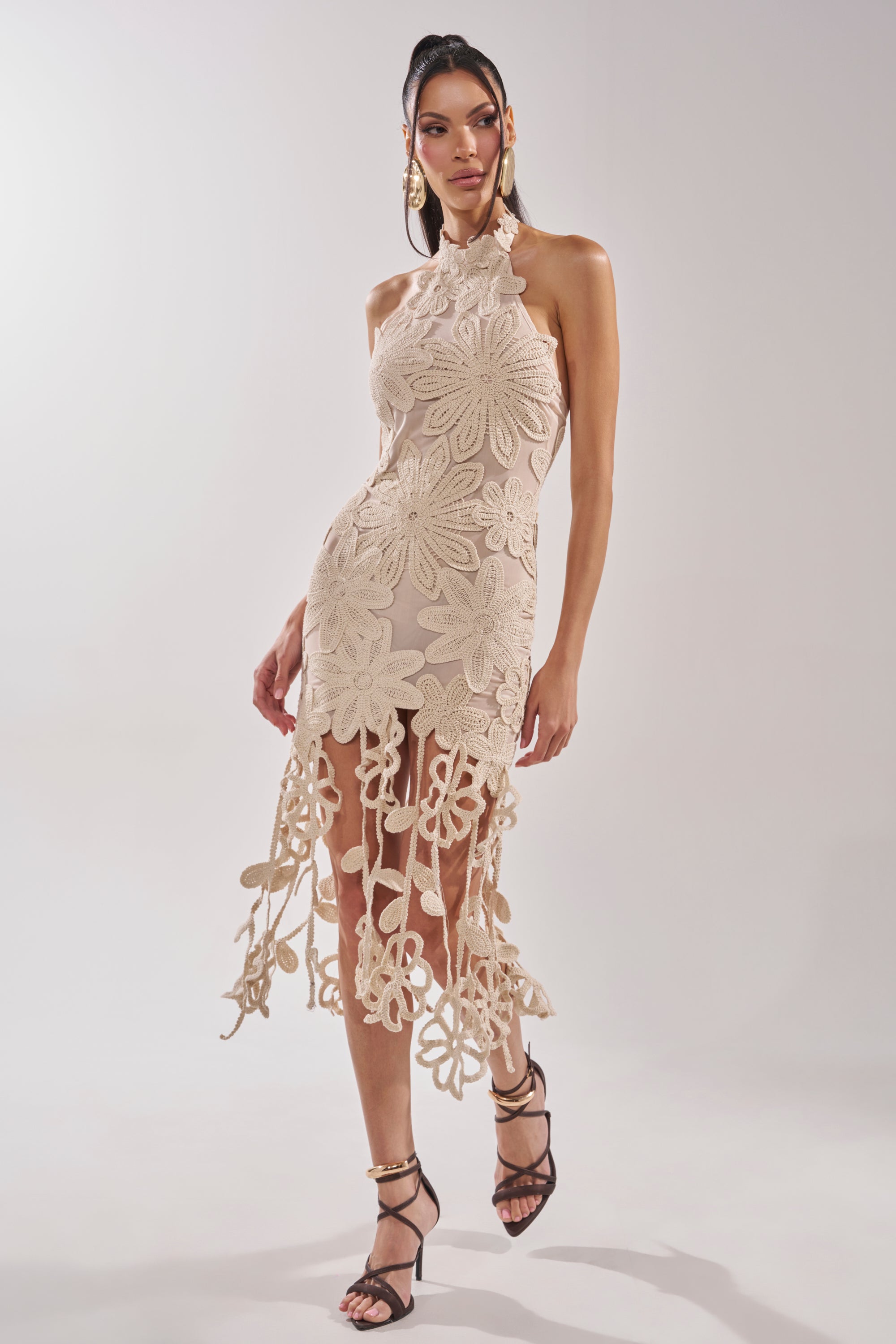A woman models the CELINE FLORAL MIDI DRESS, an ivory crochet lace dress with large floral appliqués and cut-out flower details, styled with black strappy heels and statement earrings—a stunning bridal shower choice. 1