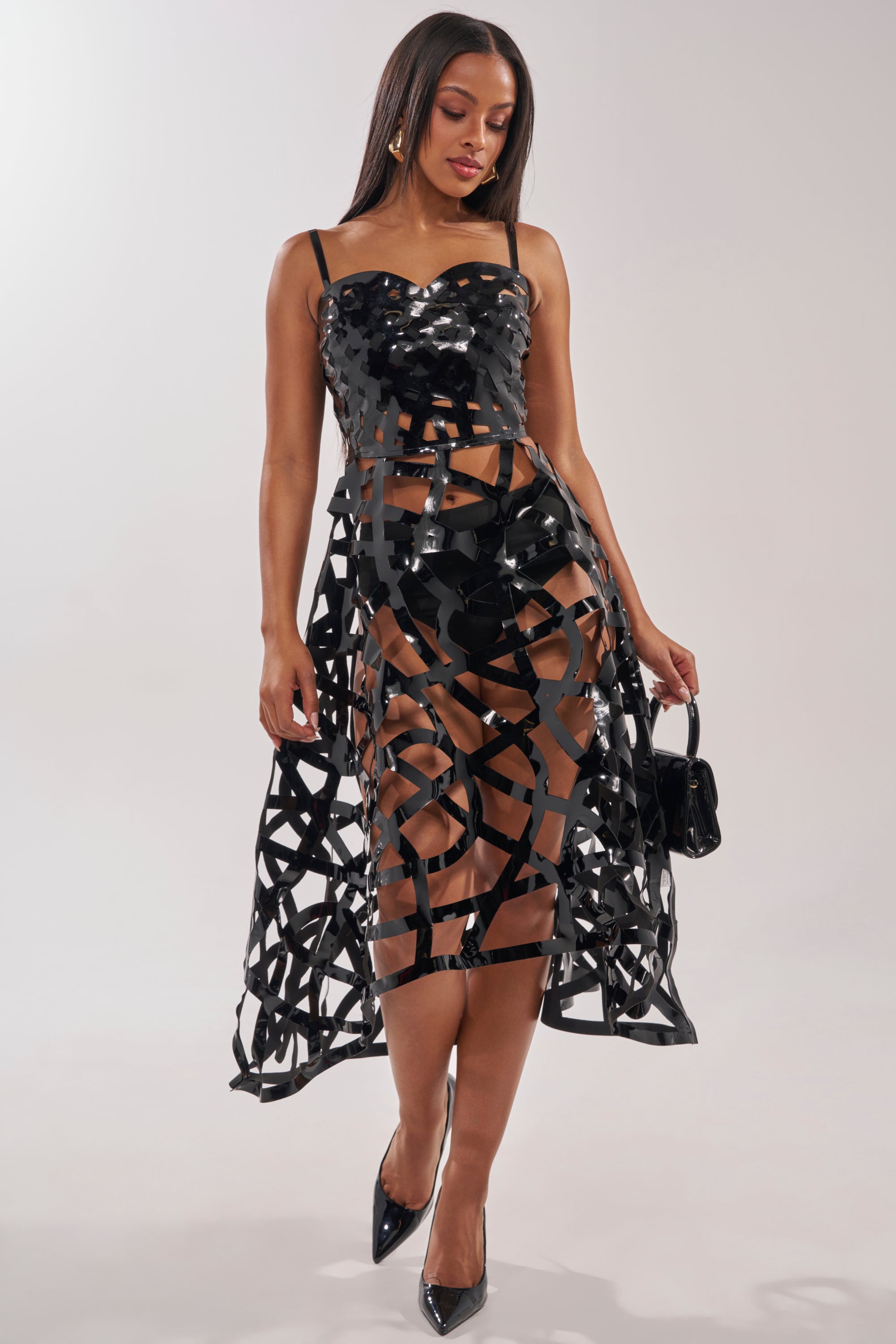 A woman models the CAUGHT YOU SLIPPING CAGED MIDI DRESS, a black strappy cutout design that shows her legs. She accessorizes with a small black purse and heels, standing against a plain light background. 0