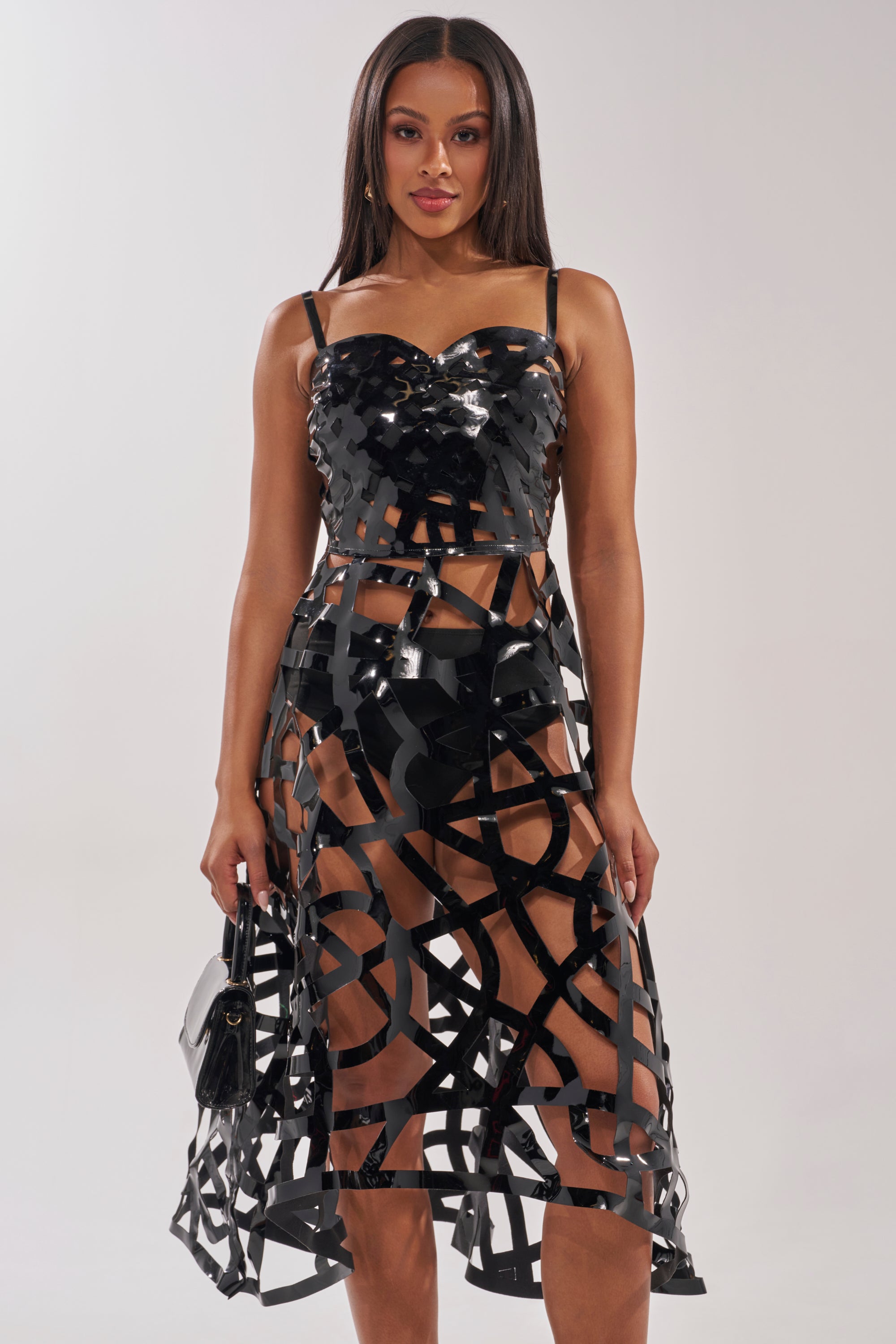 A woman stands against a plain background wearing the CAUGHT YOU SLIPPING CAGED MIDI DRESS, a black, shiny, strappy cutout dress with sheer abstract patterns, and holds a small black purse. 1