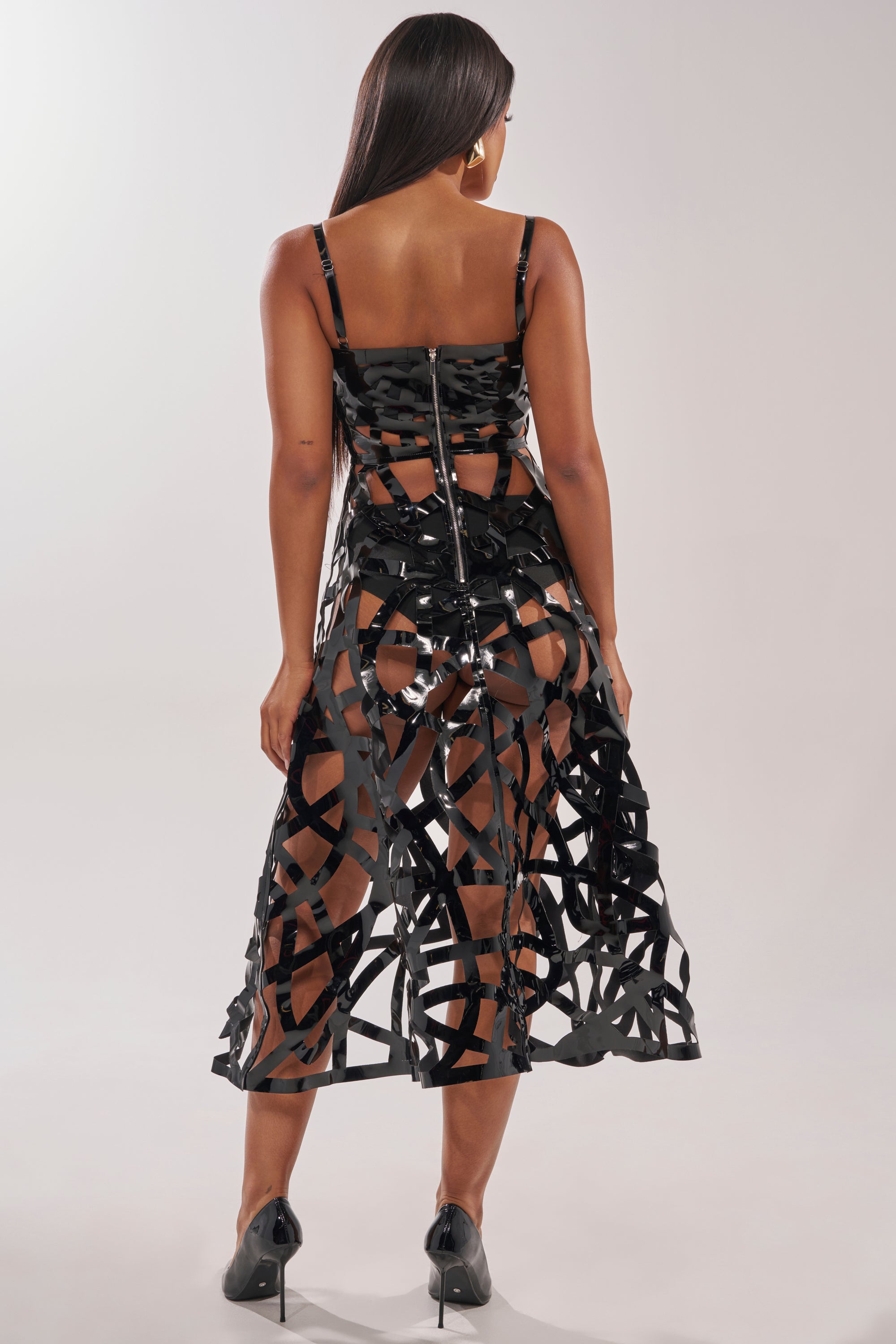 A woman faces away, wearing the CAUGHT YOU SLIPPING CAGED MIDI DRESS—a black, geometric-patterned dress with sheer panels, strappy detailing, and a visible back zipper—styled with high heels for an edgy modern look. 4