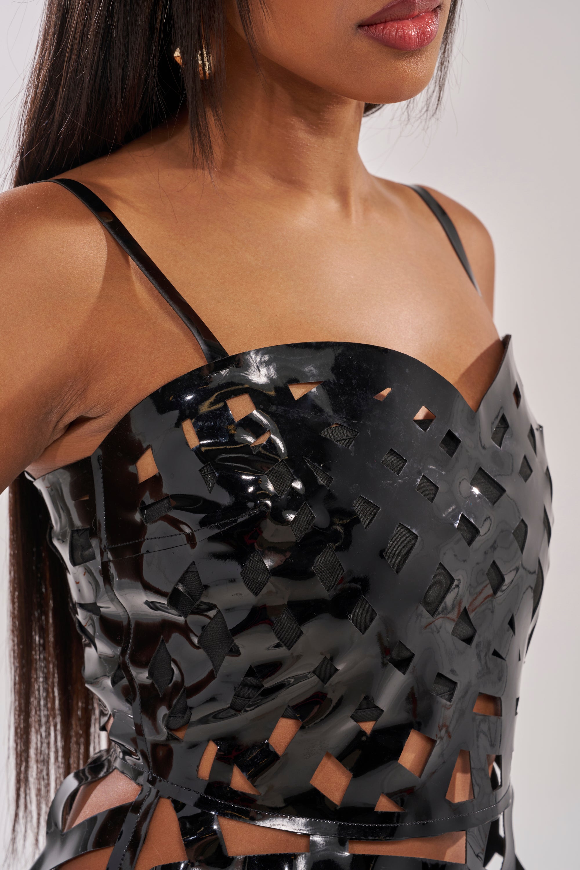 A woman models the CAUGHT YOU SLIPPING CAGED MIDI DRESS—a shiny black piece with thin straps and cutout cage details, showcasing its geometric pattern and glossy texture. Only her torso and part of her face are visible in the close-up. 5