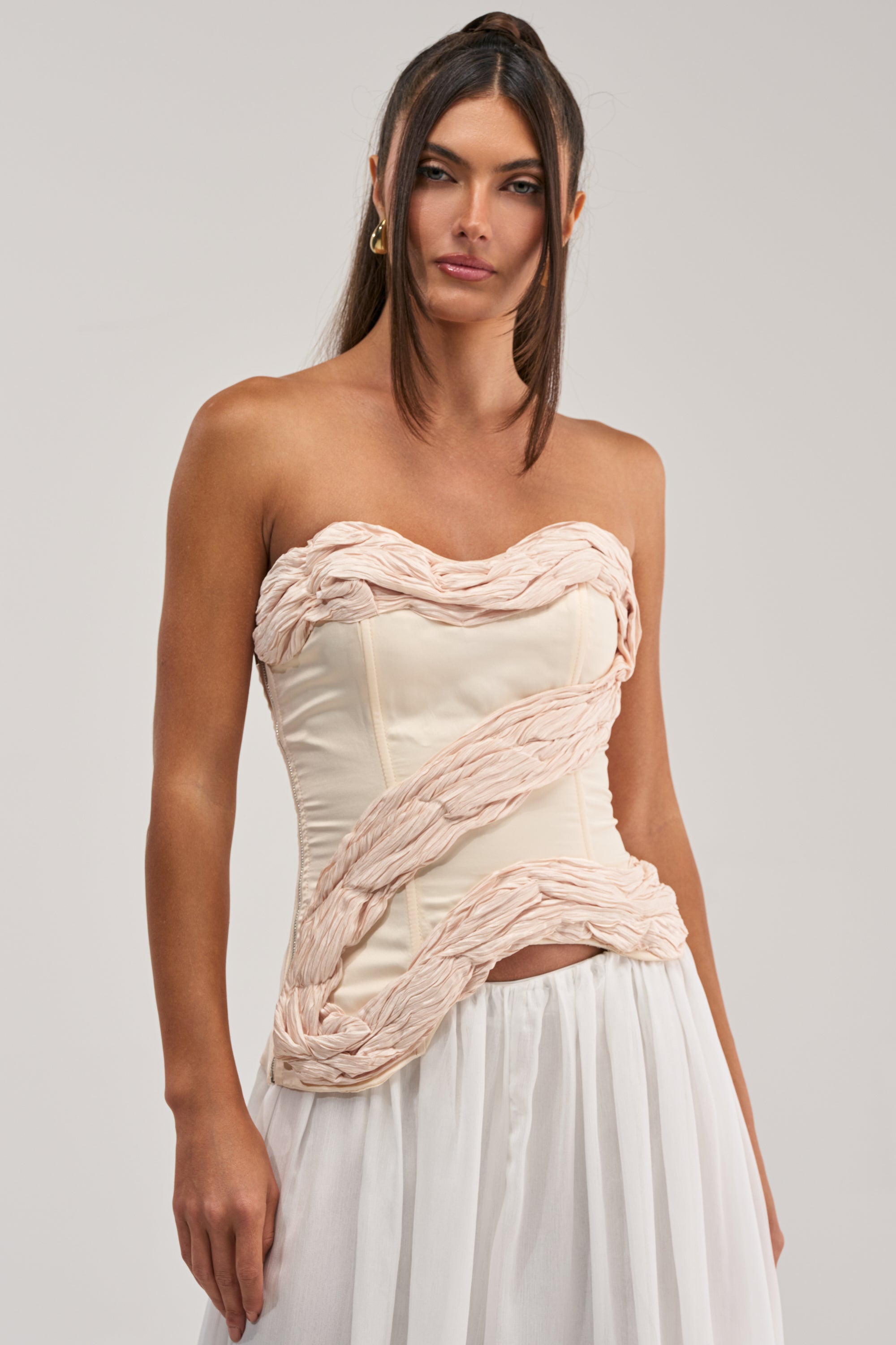 A woman models the WHITE HOT SATIN CORSET, a strapless cream top with a sweetheart neckline, twisted textured details, and a flowing white skirt, posed against a plain light background.