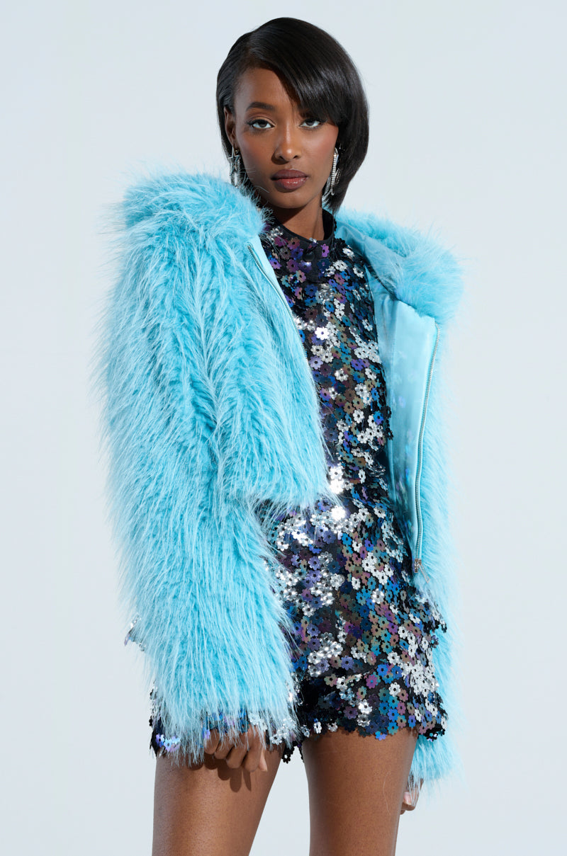 A person showcasing the vibrant KINGSTON FAUX FUR JACKET in a light blue hue, layered over a dazzling multicolored sequined dress. Their straight, shoulder-length hair complements the look as they pose against a simple light backdrop.