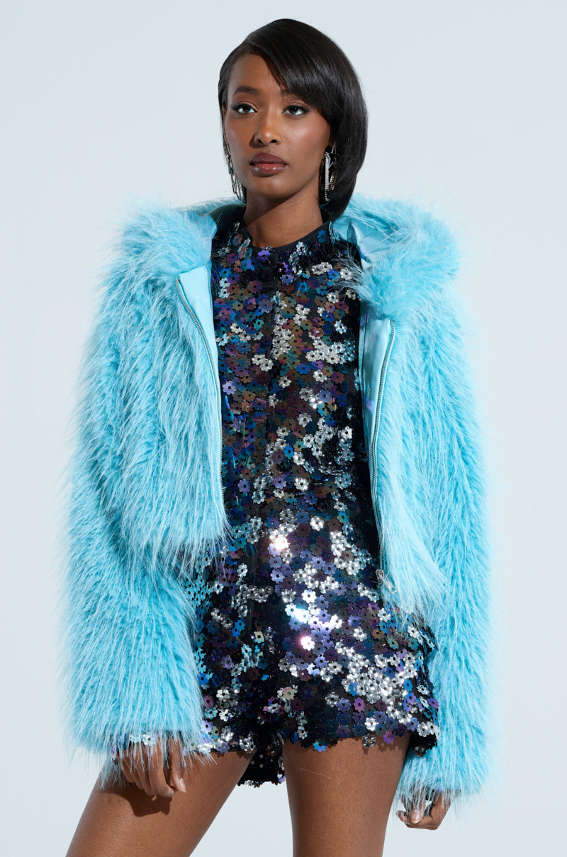 A person wearing a sparkly, multicolored sequin dress and the KINGSTON FAUX FUR JACKET stands confidently. Their hair is styled neatly, and they are adorned with large hoop earrings. The background is a light, neutral color, highlighting the vibrant AKIRA Label ensemble.