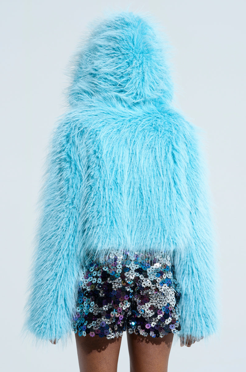 A person wearing the KINGSTON FAUX FUR JACKET in light blue with a hood stands facing away. They are also sporting striking black sequined shorts with silver and purple sparkles against a plain background, highlighting an eye-catching style perfect for any AKIRA Label enthusiast.