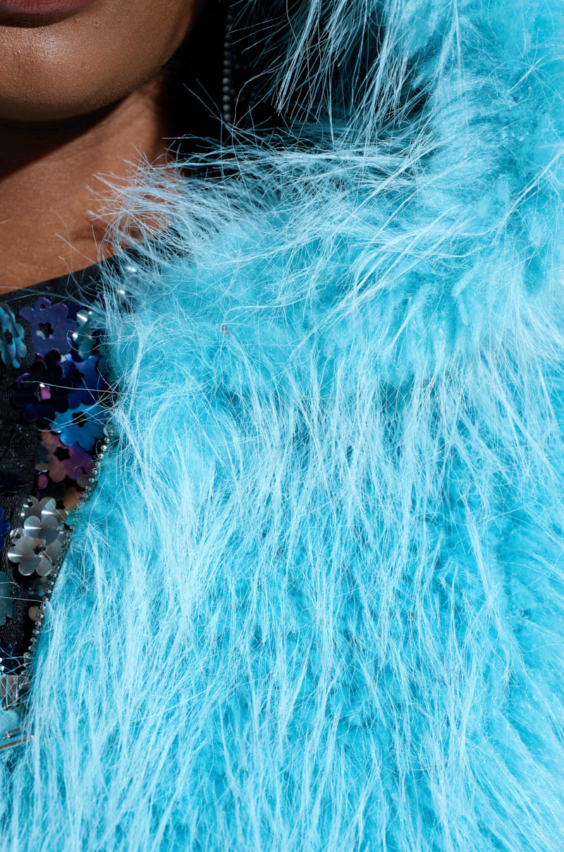 Close-up of a person wearing the vibrant aqua blue KINGSTON FAUX FUR JACKET from the AKIRA Label. Beneath it, a garment adorned with circular, multicolored sequins is partially visible, capturing a sense of texture and bold fashion.