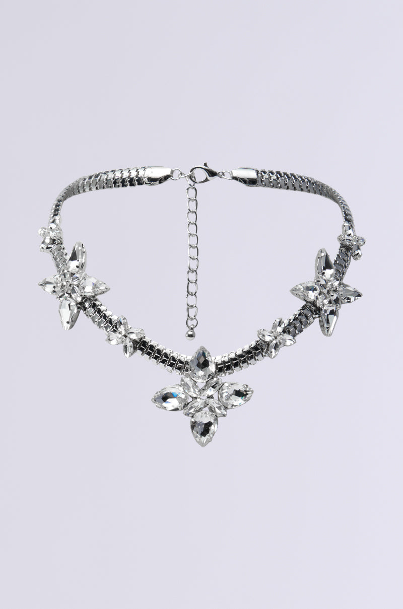 The HILLSIDE BLING NECKLACE showcases a silver herringbone-style chain adorned with three ornate, flower-shaped crystal embellishments, beautifully contrasted against a plain grey background. It is designed with an adjustable clasp to ensure a perfect fit. 0