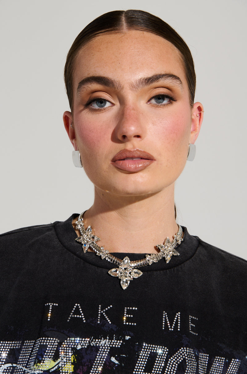 A person with slicked-back hair, wearing a black shirt featuring the text "TAKE ME," showcases the dazzling HILLSIDE BLING NECKLACE, which is decorated with starburst rhinestone charms. Their bold eye makeup and neutral lipstick complement the neutral background behind them. 1