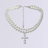 The PEARL DREAMING CROSS CHOKER is an exquisite three-strand necklace adorned with pearly beads and a hanging cross pendant. This layered accessory features a silver chain clasp for adjustable fitting, beautifully contrasted against a plain light gray background.