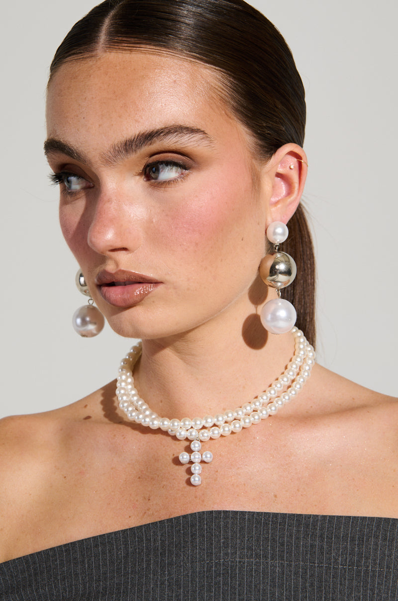 A woman with long brown hair wears the PEARL DREAMING CROSS CHOKER and large pearl earrings, paired with a strapless dark pinstriped outfit. The backdrop is a simple gray. 5