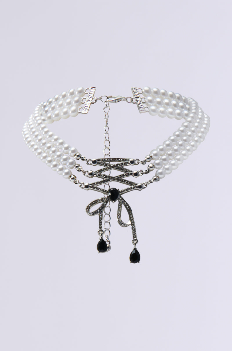 The COQUETTE DREAMING CHOKER is an elegant accessory comprising three strands of pearl beads, accentuated by a central black stone and a delicate bow design adorned with rhinestones. Its adjustable chain closure enhances its refined appearance, making it stand out against a simple backdrop.