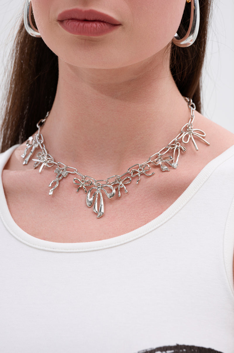 A woman with straight brown hair wears the CUTESY NECKLACE, featuring bow-shaped charms and an oversized link chain, paired with large silver hoop earrings and a white top.