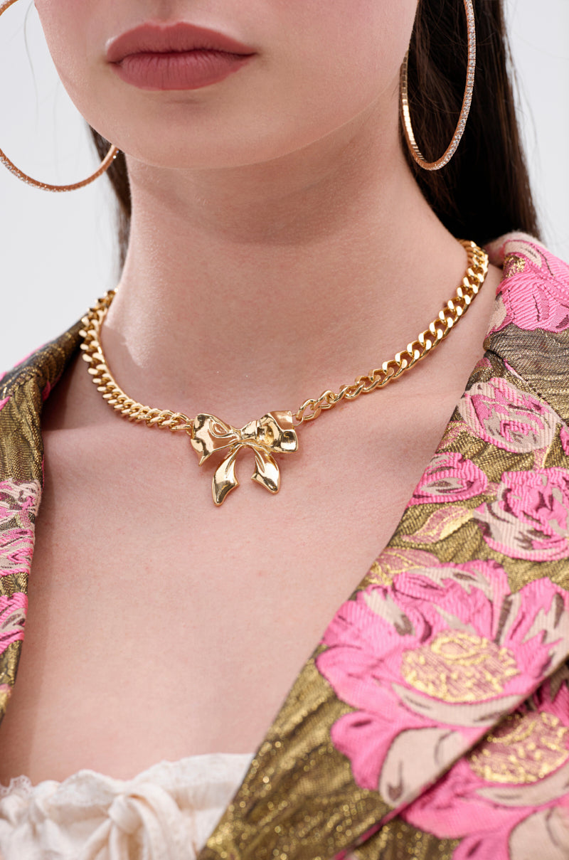 A woman wears large hoop earrings and the BELLA NECKLACE, a chunky curb link design with a bow pendant. She is dressed in a floral-patterned jacket featuring pink and gold accents. The image is cropped to her lower face and upper torso.