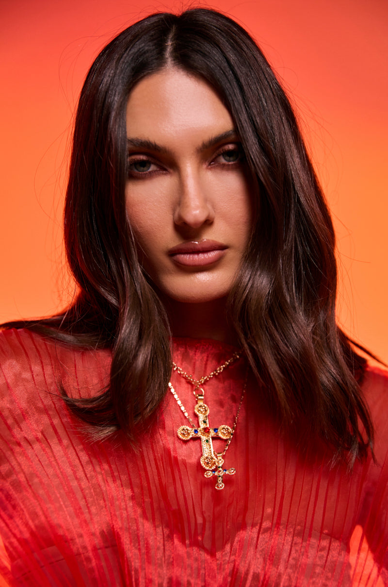 A woman with long brown hair wears a sheer red pleated top and the LAYERED UP NECKLACE, posing against a vibrant orange background.