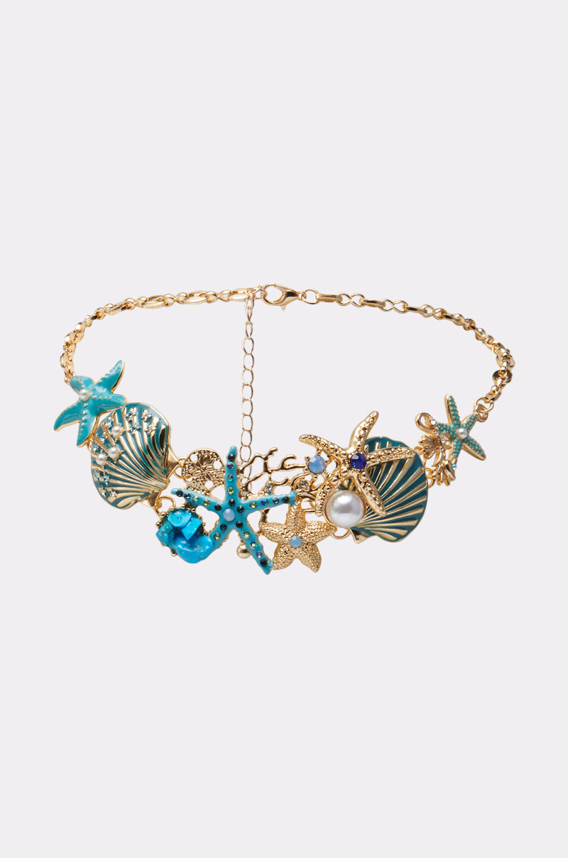 The TO THE SHORE NECKLACE is a gold chain with coastal glam, adorned with sea-inspired charms—blue enamel starfish, gold seashells, a pearl, and ocean motifs—shown on a plain white background.