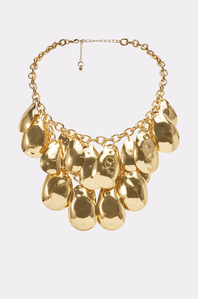 The ON A TRIP NECKLACE is a chunky gold chain with large, shiny, irregular pendants—including hammered teardrop shapes—hanging from a thick, adjustable choker clasp.