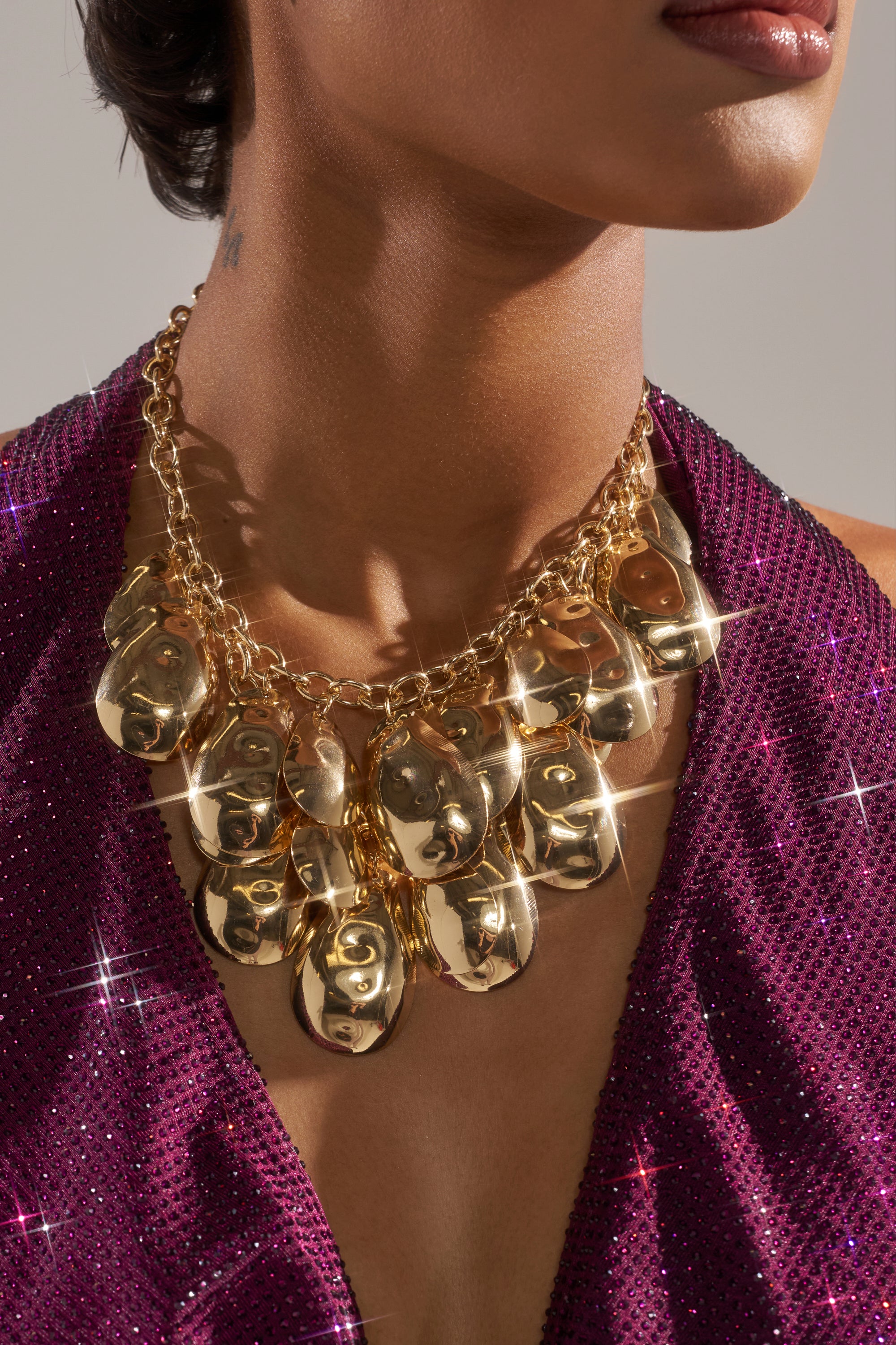 A woman wears a deep purple, sparkly halter top and the ON A TRIP NECKLACE—a chunky gold choker featuring hammered metal mask-shaped pendants. Her jewelry catches and reflects the light, adding sparkle to the image.