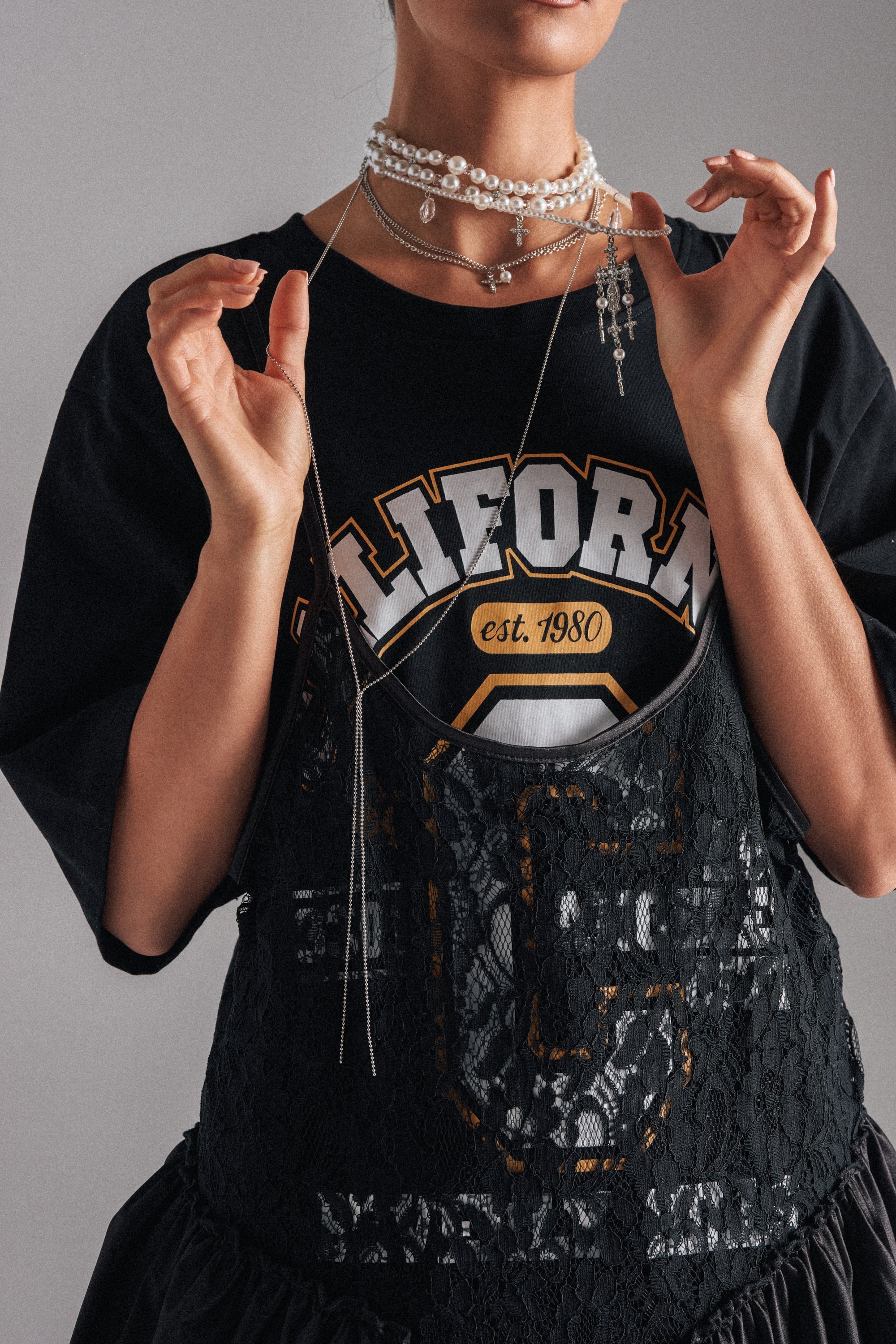 A woman wears the HERE FOR THE VIBES NECKLACE layered with other necklaces, styled with a black lace dress and a "California est. 1980" shirt, shown from shoulders to waist against a plain gray background. 0