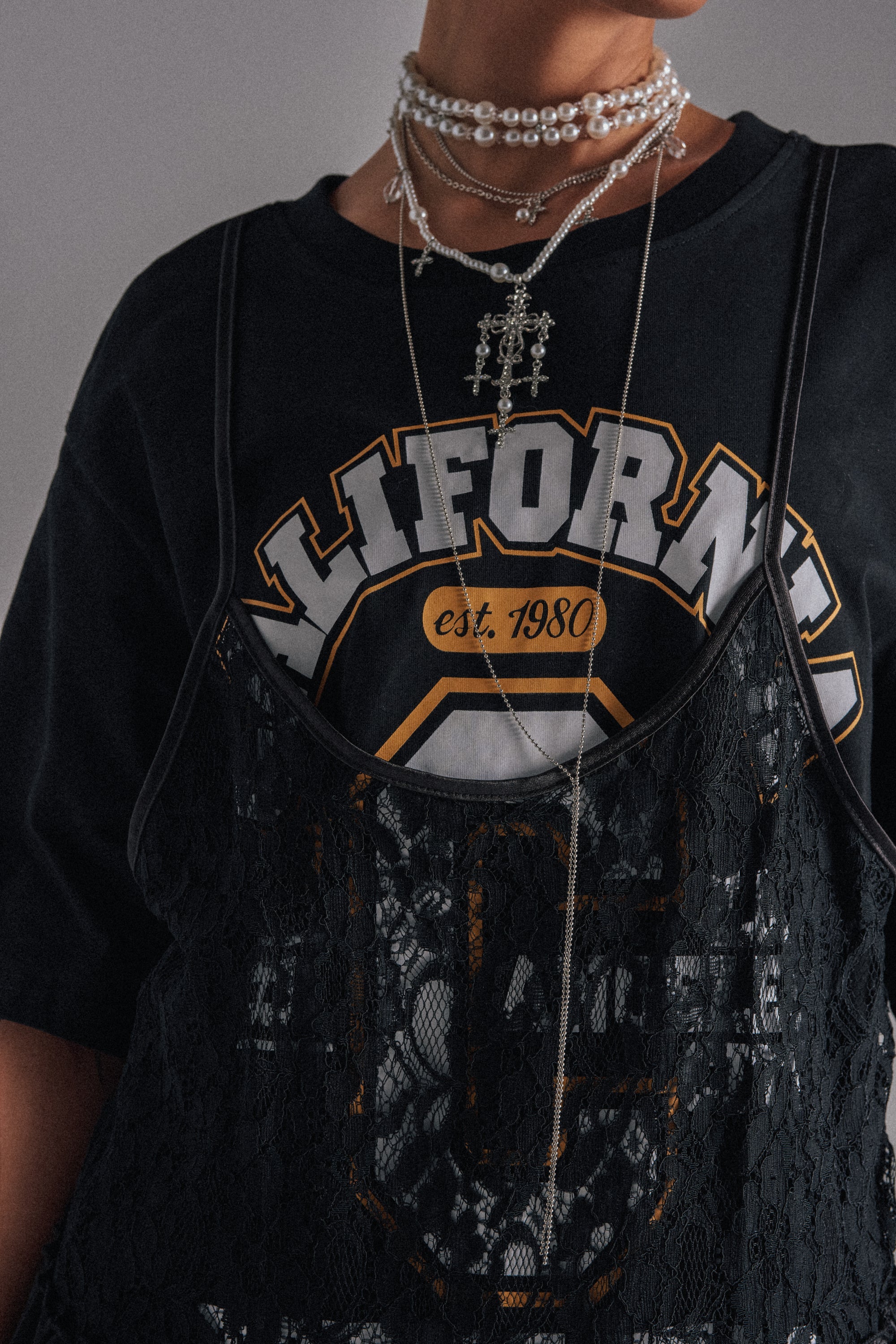 A person wears a black "California est. 1980" t-shirt, layered with a black lace camisole and the HERE FOR THE VIBES NECKLACE, featuring chunky pearls and an embellished cross pendant. The image is cropped at the shoulders. 1