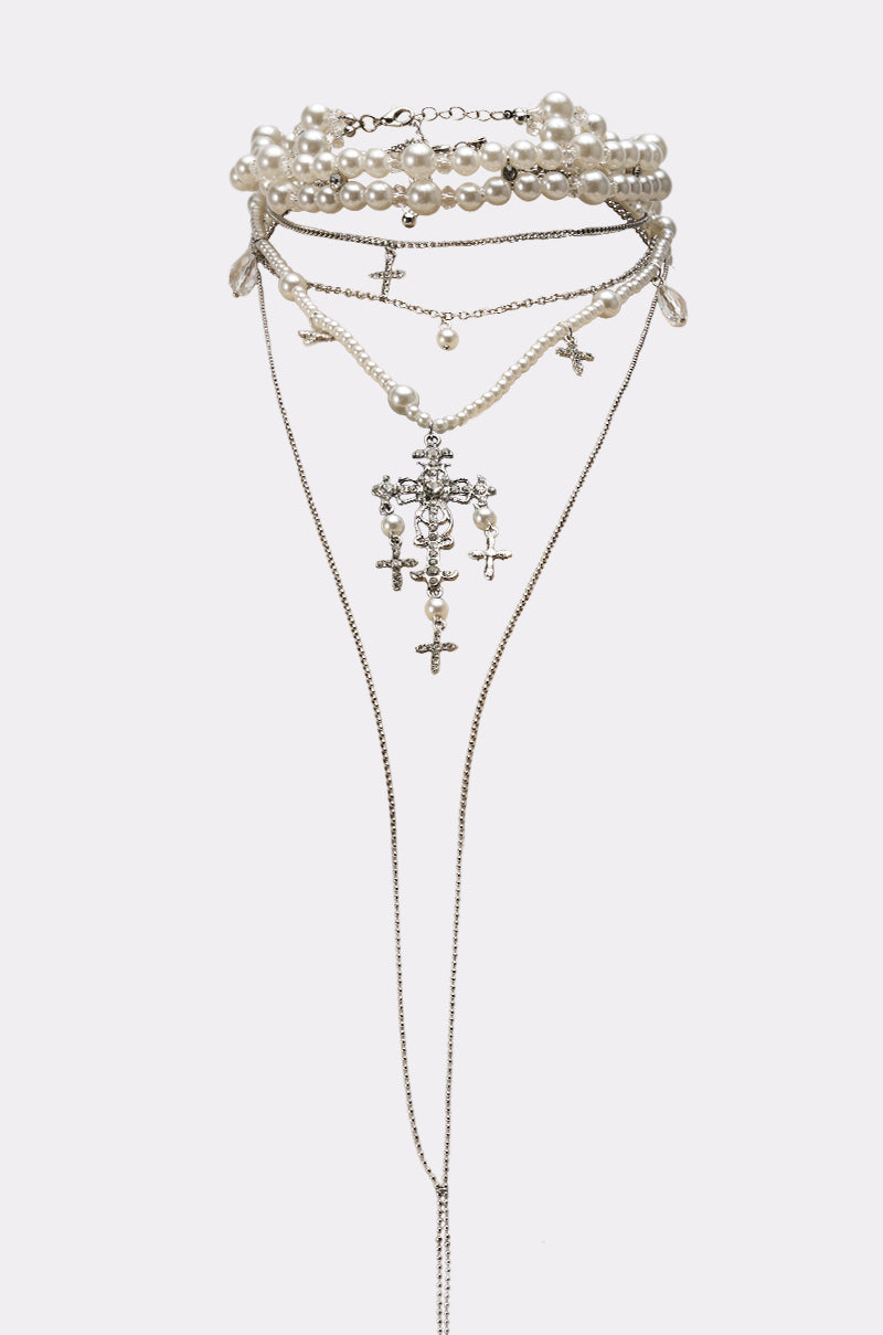 The HERE FOR THE VIBES NECKLACE is a layered chain design with faux pearl strands, small crosses, and decorative charms. Long, delicate chains drape elegantly, creating an ornate look against a plain white background.