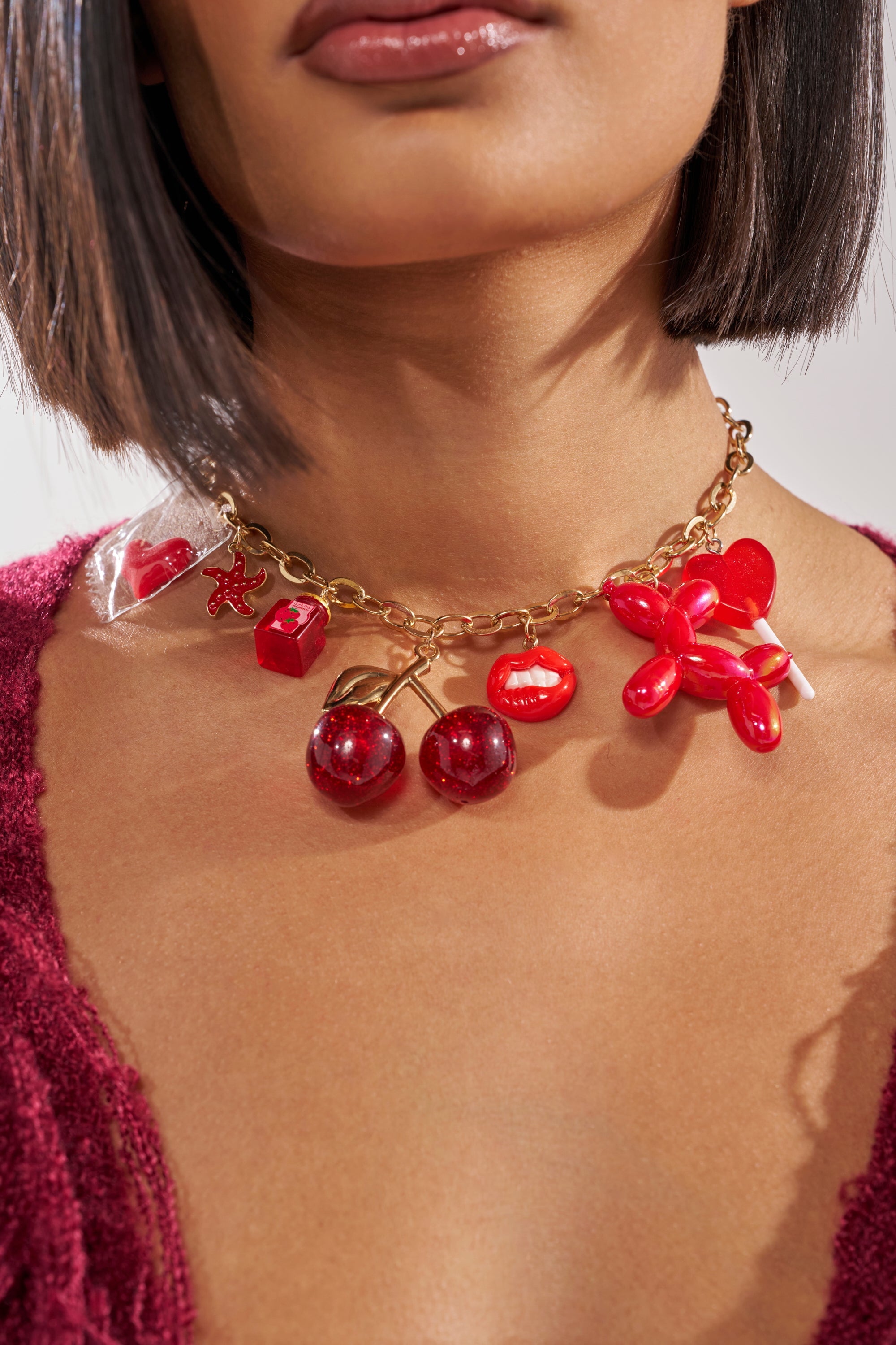 A woman wears the HAVE SOME FUN NECKLACE—a chunky gold-tone Y2K chain with red charms (cherries, heart, wrapped candy, balloon dog)—perfectly paired with a textured burgundy top. 1