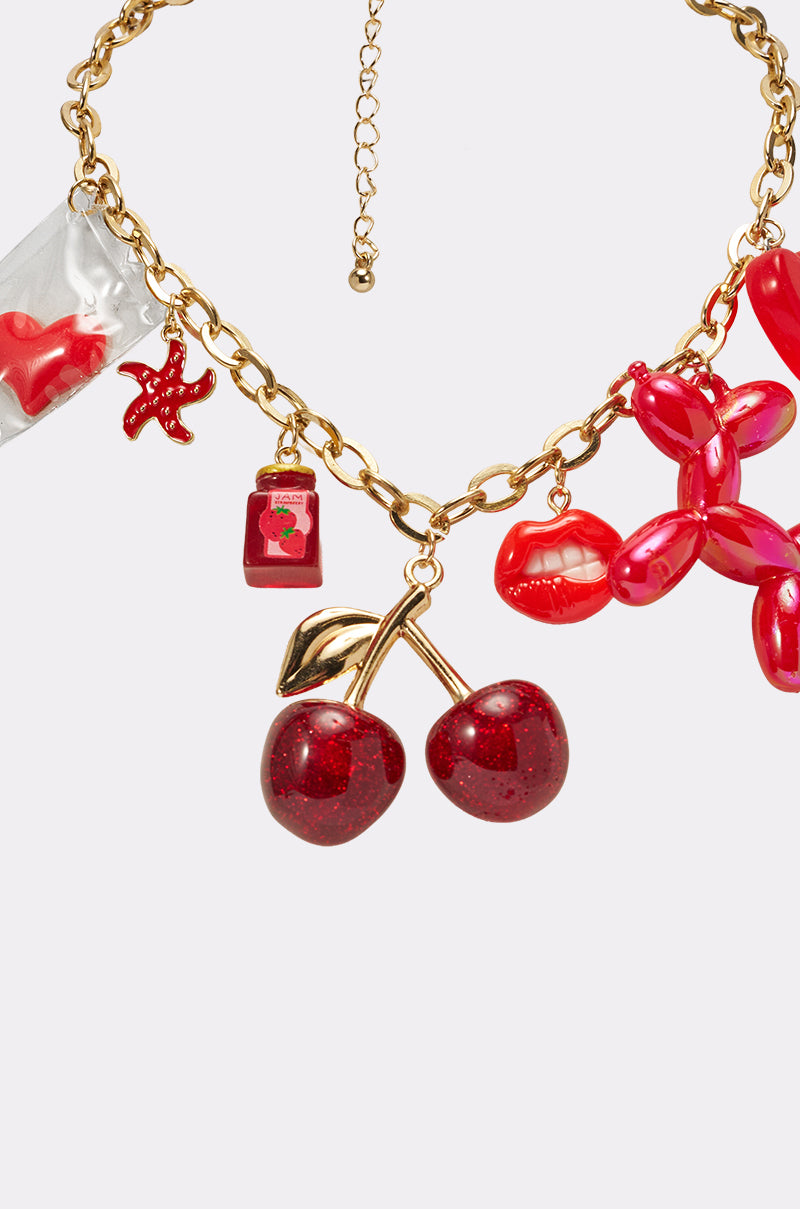 The HAVE SOME FUN NECKLACE is a chunky gold chain with playful red charms—sparkly cherries, a balloon dog, lips, a tiny bottle, starfish, and a toy—channeling Y2K vibes against a white background. 3