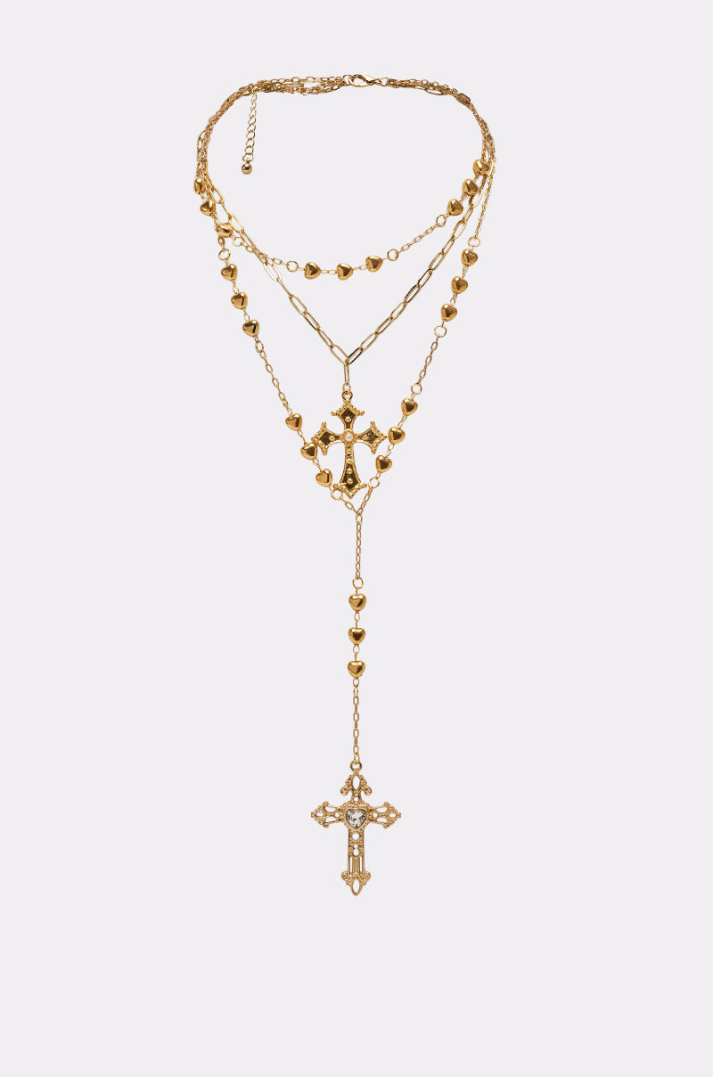 The DRIPPED OUT CROSS LARIAT is a layered gold necklace with small heart charms and a cross pendant, shown against a light gray background. 0