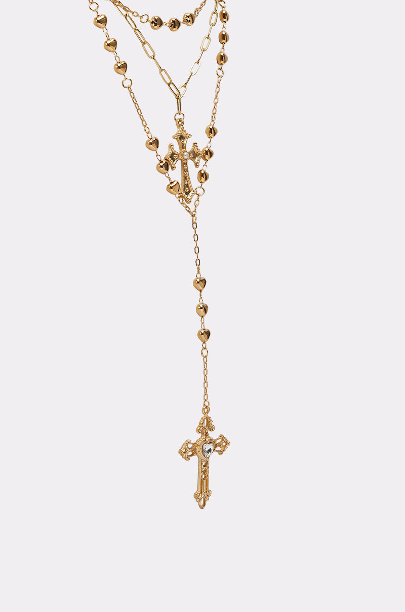 The DRIPPED OUT CROSS LARIAT is a gold-tone layered necklace with ornate cross pendants, decorative beads, and heart accents along the chains, showcased against a simple light background. 3