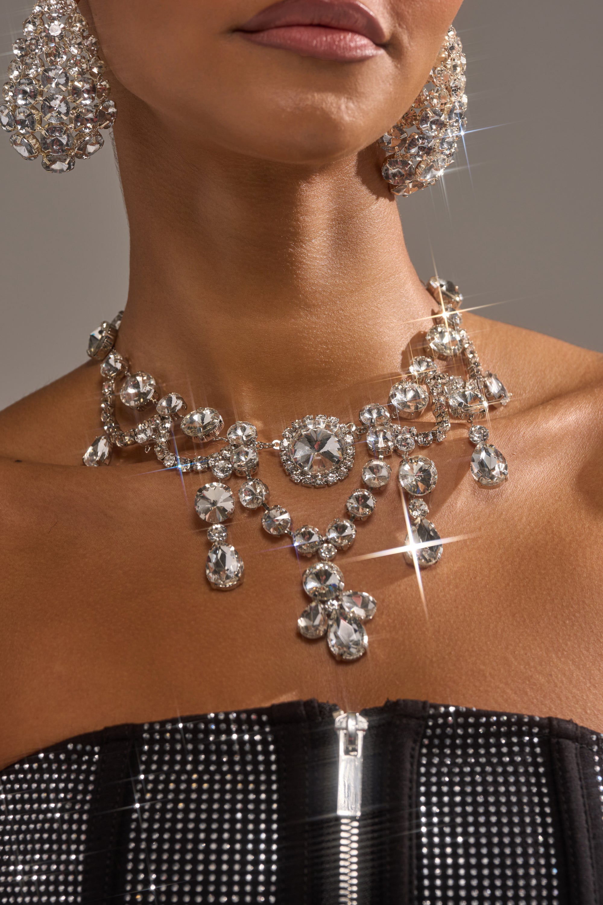 A woman wears the YOU LOVE ME BLING NECKLACE and large sparkling earrings, paired with a black strapless rhinestone-studded top with a visible silver zipper.
