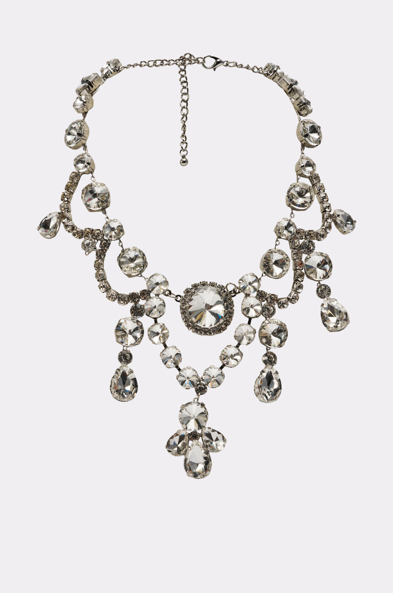 The YOU LOVE ME BLING NECKLACE is a silver statement piece with large clear faceted gemstones, ornate symmetry, dangling crystal accents, and an adjustable chain clasp—perfect for true Hollywood glamour.