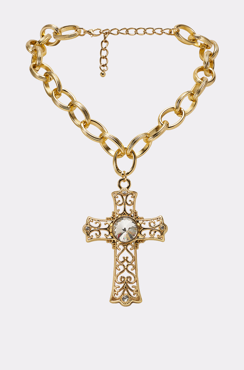 The ALL MY LOVE NECKLACE is a chunky, oversized gold necklace with an ornate cross pendant, filigree detailing, and a large clear gemstone at the center, displayed on a plain light background. 0