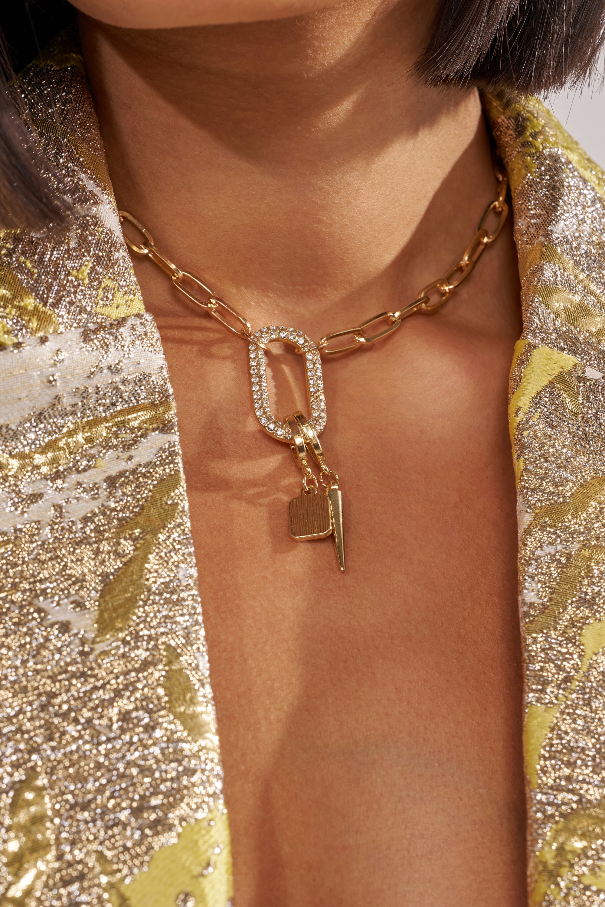A close-up of a person wearing the DESIGNER NECKLACE, a gold-tone paperclip chain with a rhinestone-studded oval link and two gold charms, paired with a shimmery gold and silver patterned jacket. 1