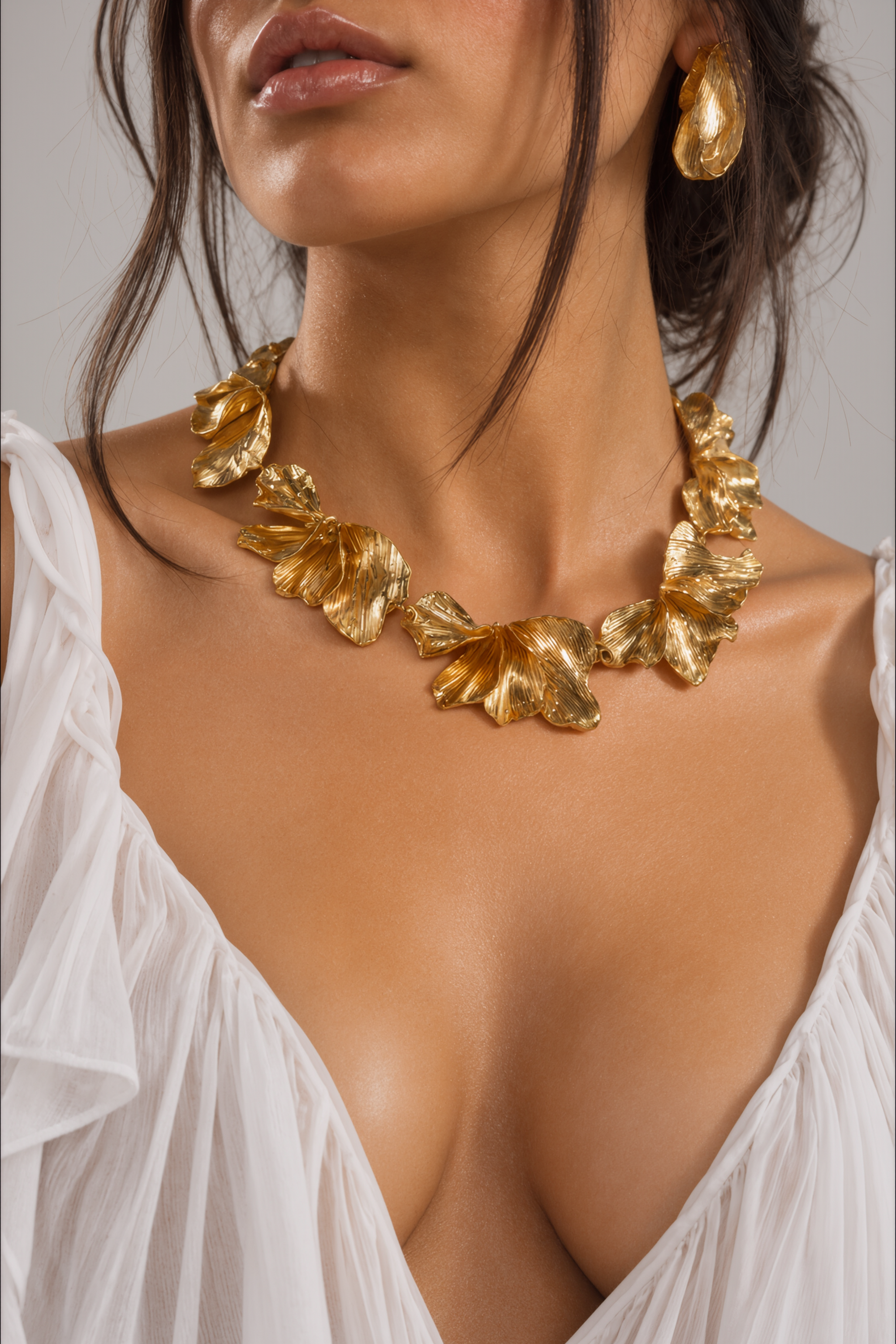 A woman wearing a white, low-cut top showcases the bold, oversized ON A BEACH NECKLACE paired with matching gold earrings. Her loosely styled hair partially covers her face.