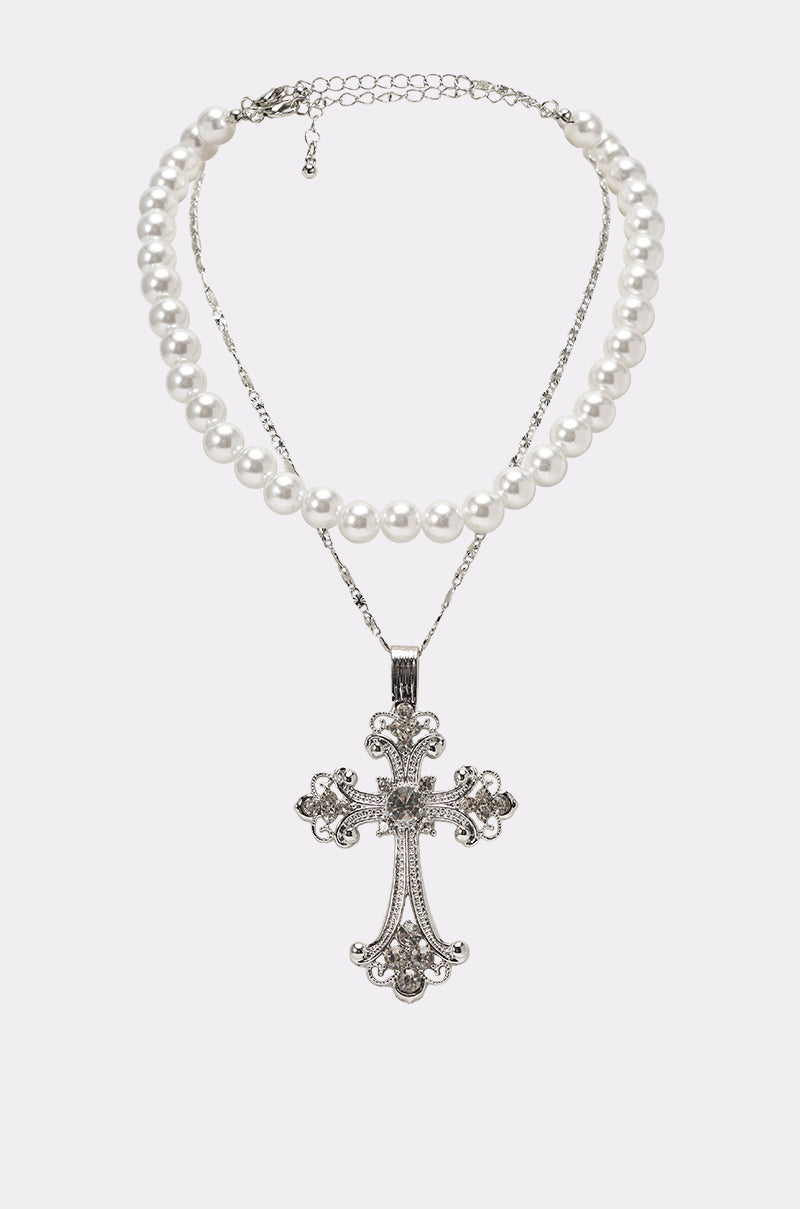 The REGENCY NECKLACE is a two-piece set featuring a faux pearl choker and a delicate silver chain, both joined by an ornate silver cross with intricate designs and sparkling jewels. 0