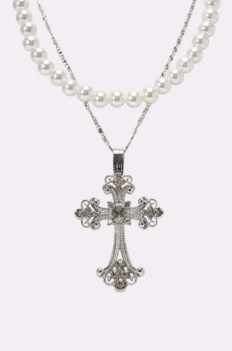 The REGENCY NECKLACE features a silver ornate cross pendant on a delicate chain, layered with white faux pearls, set against a plain light background. 3
