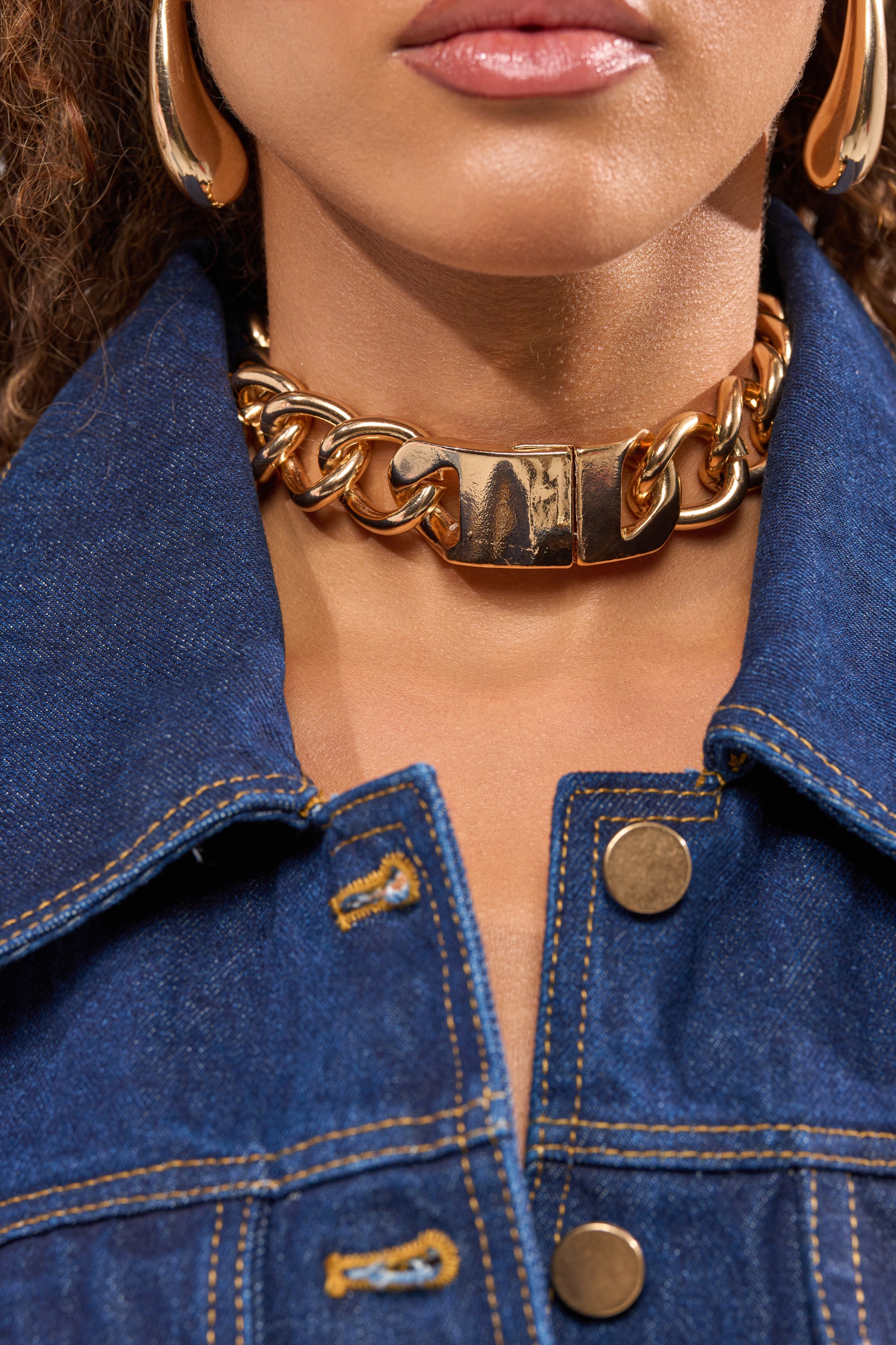 A close-up highlights the ITS HEAVY NECKLACE IN GOLD—a bold streetwear choker—styled with large gold hoop earrings and a blue denim jacket with gold buttons, focusing on the wearer's neck and lower face. 1
